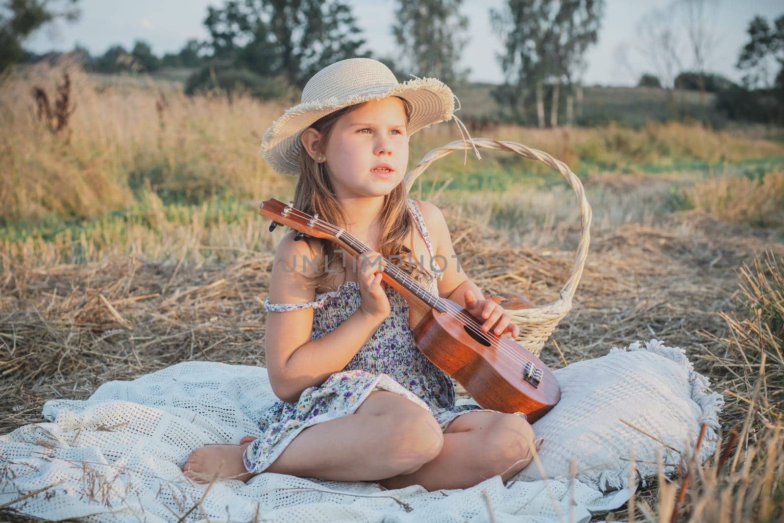 Pretty little girl play ukulele sitting on blanket on field. Portrait of child in panama hat looking away. Outdoor walking and picnic. Music instrument, hobby. Summer vacation, countryside lifestyle.
