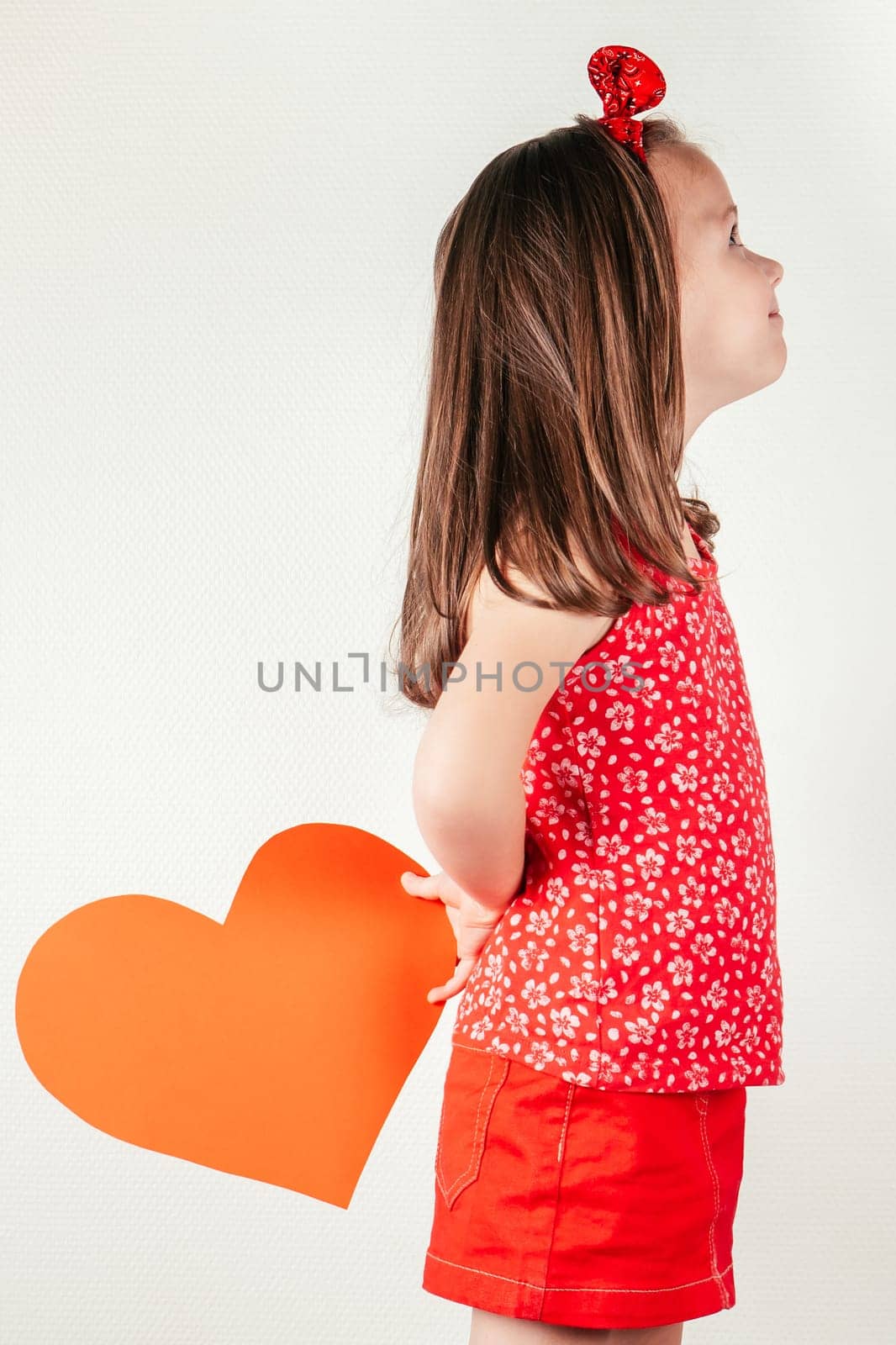 Smiling little girl hide red heart shaped greeting card behind back. Portrait of child on white, side view. Give paper cut valentine. Saint Valentine's Day celebration, holiday of all lovers.