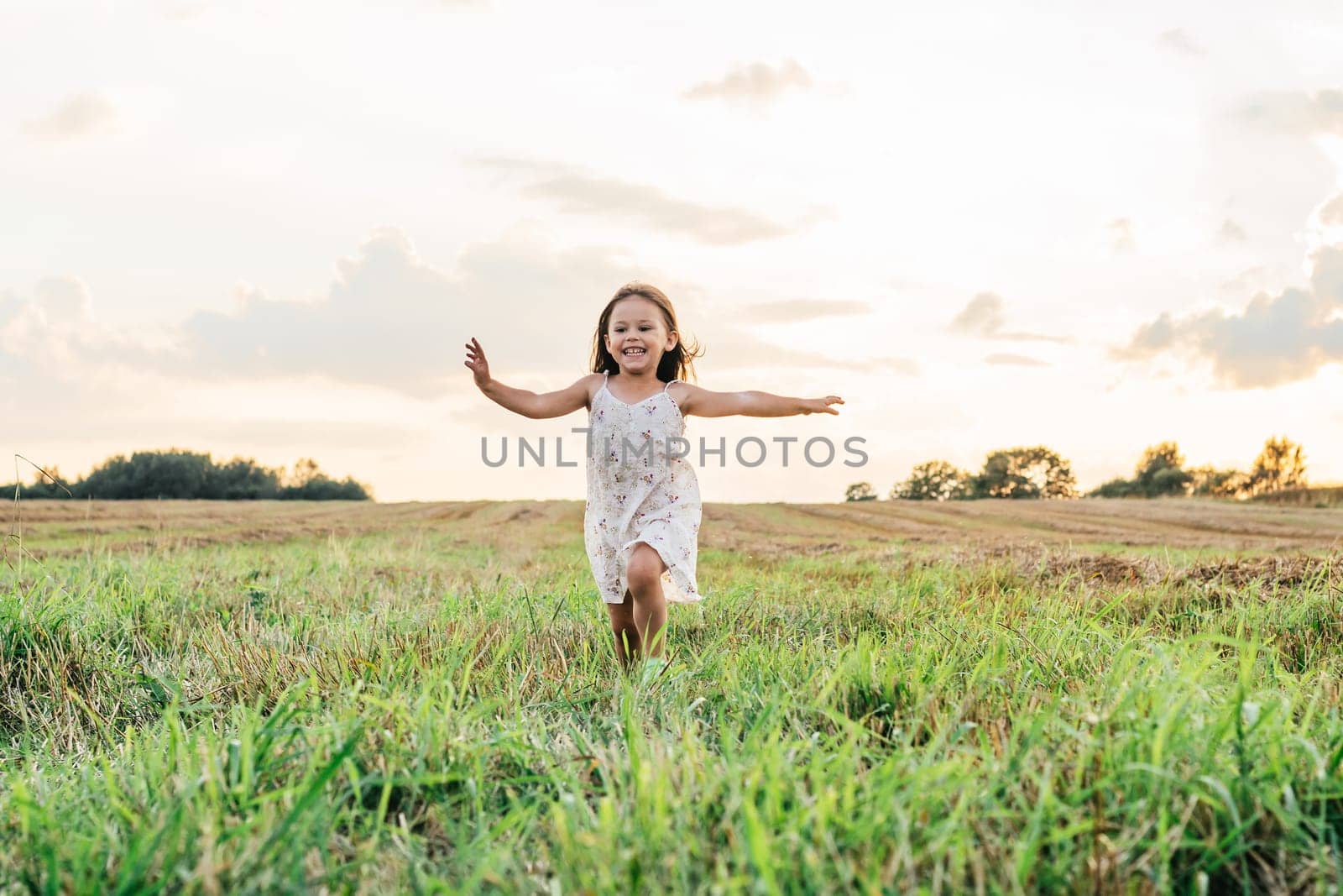 Smiling little girl joyfully running across field. Happy carefree child on grassy meadow. Outdoor walking. Beautiful sunny sky. Summer vacation, countryside lifestyle. Picturesque landscape.