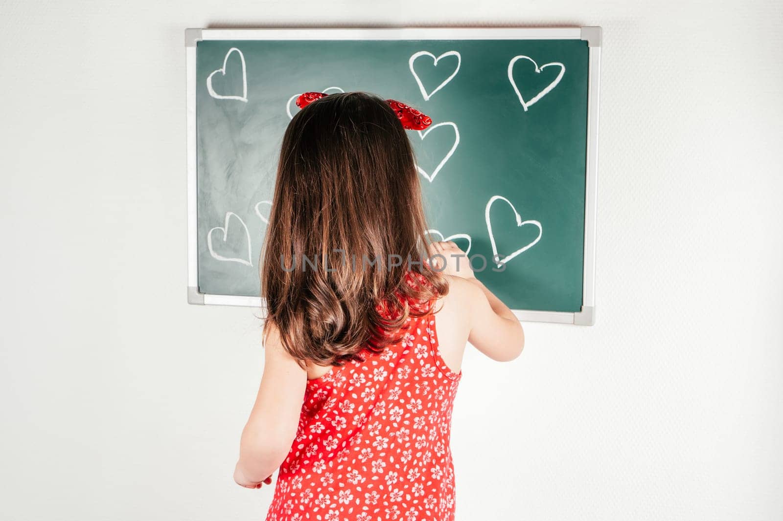 Little girl in red dress turning away draw with chalk hearts on green blackboard. Portrait of child on white, back view. Drawing, festive pattern. Saint Valentine's Day, holiday of all lovers.