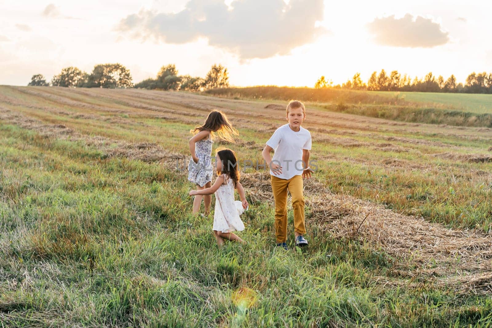 Little children run forward across field turning back. Carefree kids play together on grassy meadow. Outdoor walking. Beautiful sunny sky, sunset. Summer vacation, countryside lifestyle.