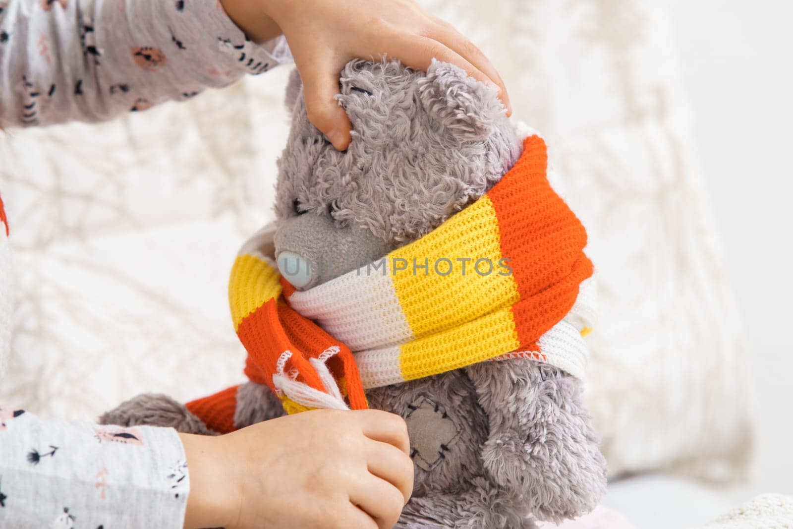 Hands of little girl tie knitted white-orange-yellow scarf around neck of sick teddy bear. Kid in sleepwear playing with stuffed toy. Disease, treatment. Doctor and patient. Closeup photo.