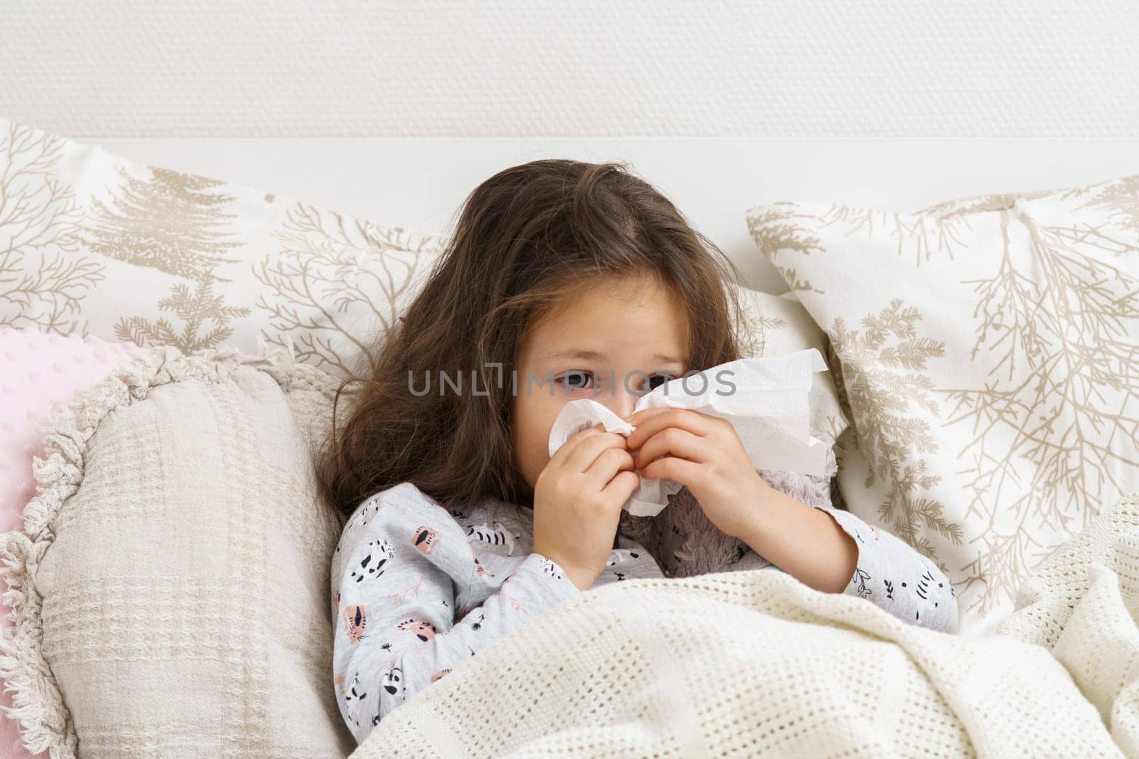 Little girl blow stuffy nose with white handkerchief. Portrait of unhealthy child. Kid in sleepwear lie under blanket and have bed rest. Home treatment. Runny nose, snot, cold symptoms, cure.