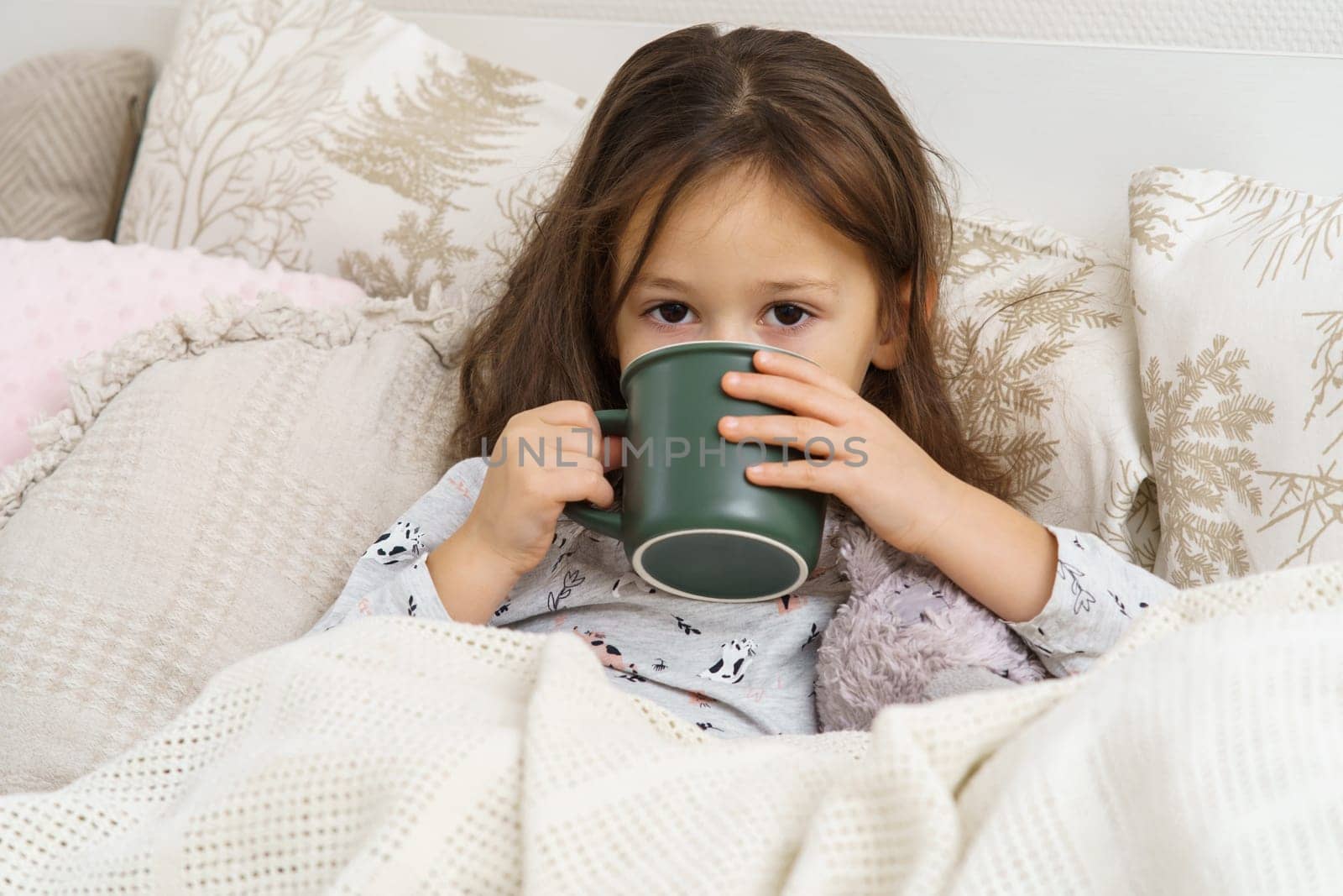 Sick little girl in sleepwear lying under blanket on bed and drink beverage from cup. Portrait of unhealthy child. Kid have bed rest. Home treatment, cold, disease. Taking medicament.