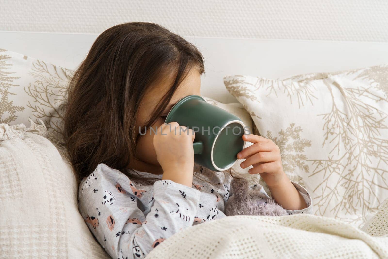 Little girl in sleepwear lie under blanket and take sip of drink covering face with mug. Portrait of unhealthy child, side view. Kid have bed rest. Home treatment, cold, disease. Taking medicament.