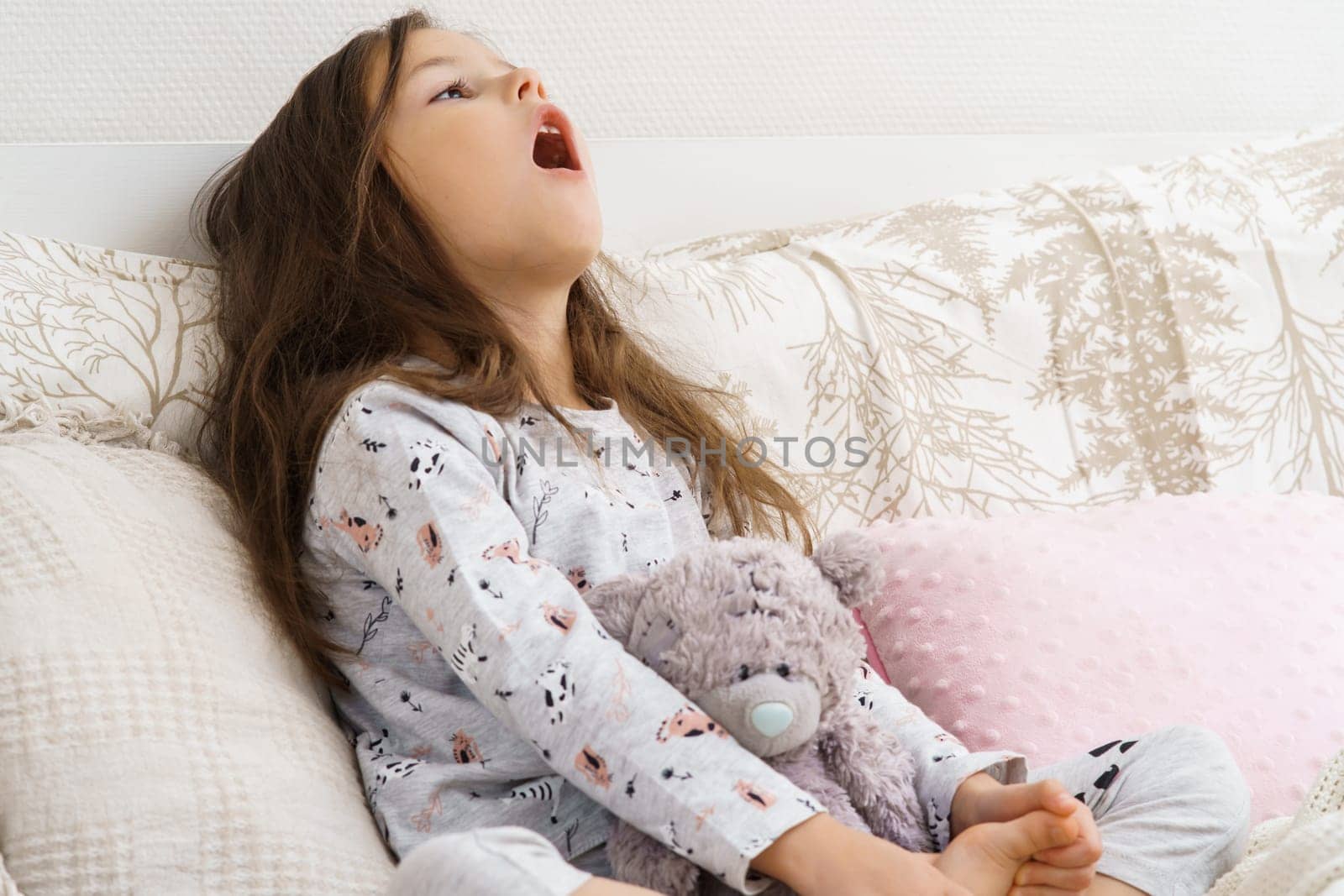 Ill little girl in sleepwear sitting on couch gargle with medicine. Portrait of child, preschooler. Kid and toy have bed rest. Sore throat, home treatment. Taking medicament, cold, disease.