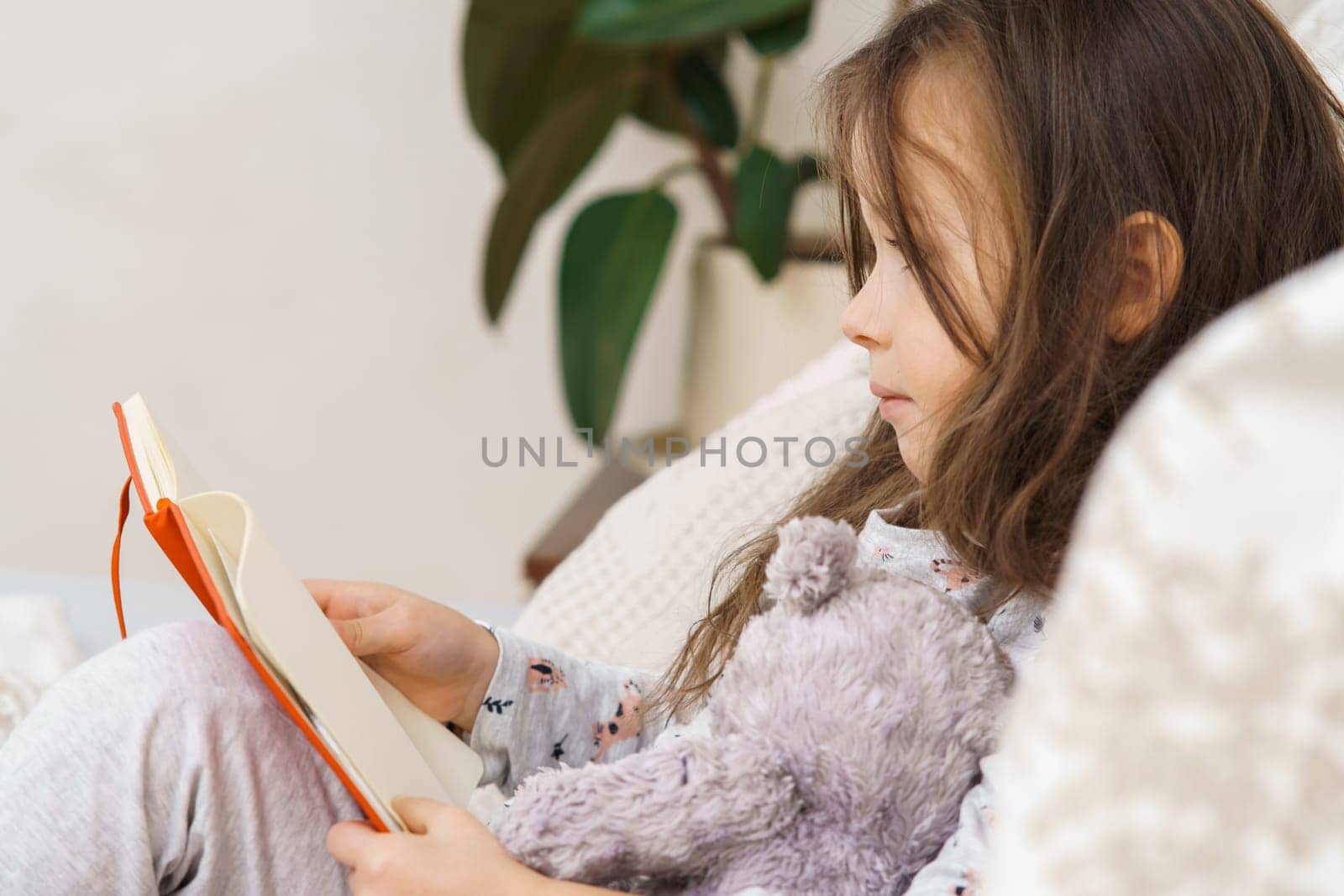 Adorable little girl in sleepwear lying on couch and reading book. Portrait of child, preschooler, side view. Kid relax at home with storybook and teddy bear. Education, hobby, lifestyle.