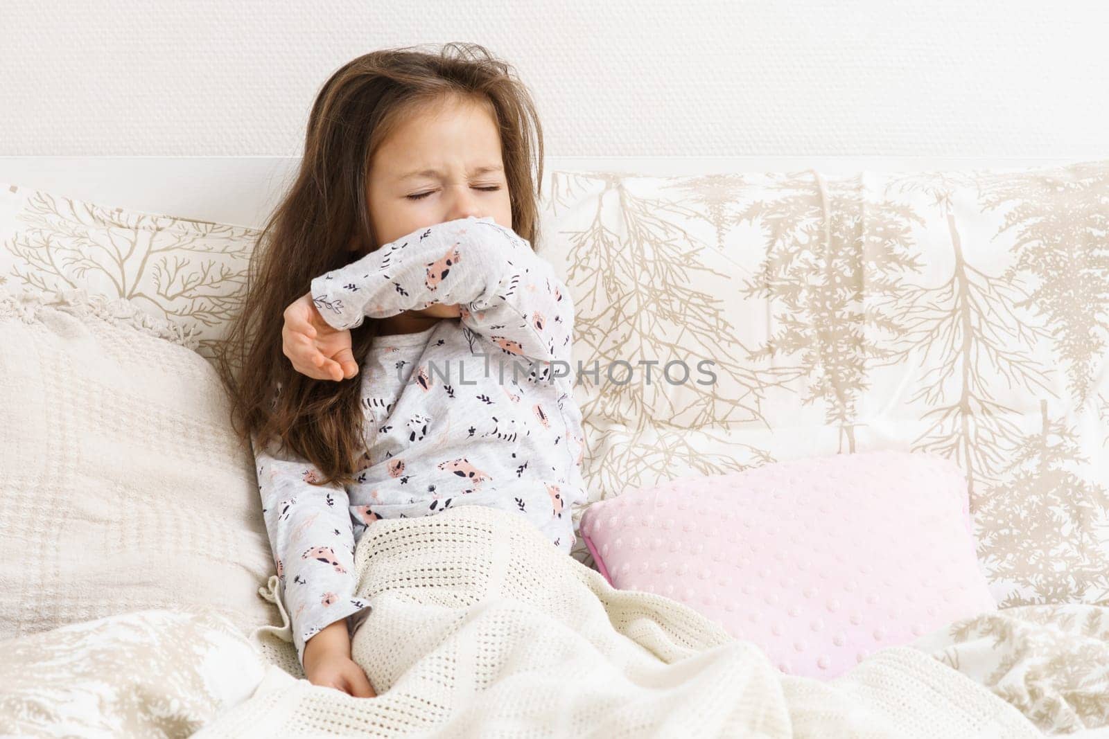 Little girl in sleepwear sit on bed under blanket and yawn with closed eyes covering mouth. Portrait of awaking child. Kid wake up at morning. Cozy bedclothes for sleeping. Resting, awakening.