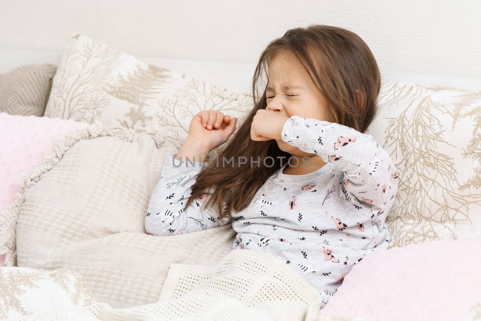 Cute little girl in sleepwear sit on bed under blanket and yawn squinting eyes. Portrait of awaking child. Kid wake up at morning. Cozy bedclothes for sleeping. Resting in bedroom, awakening.