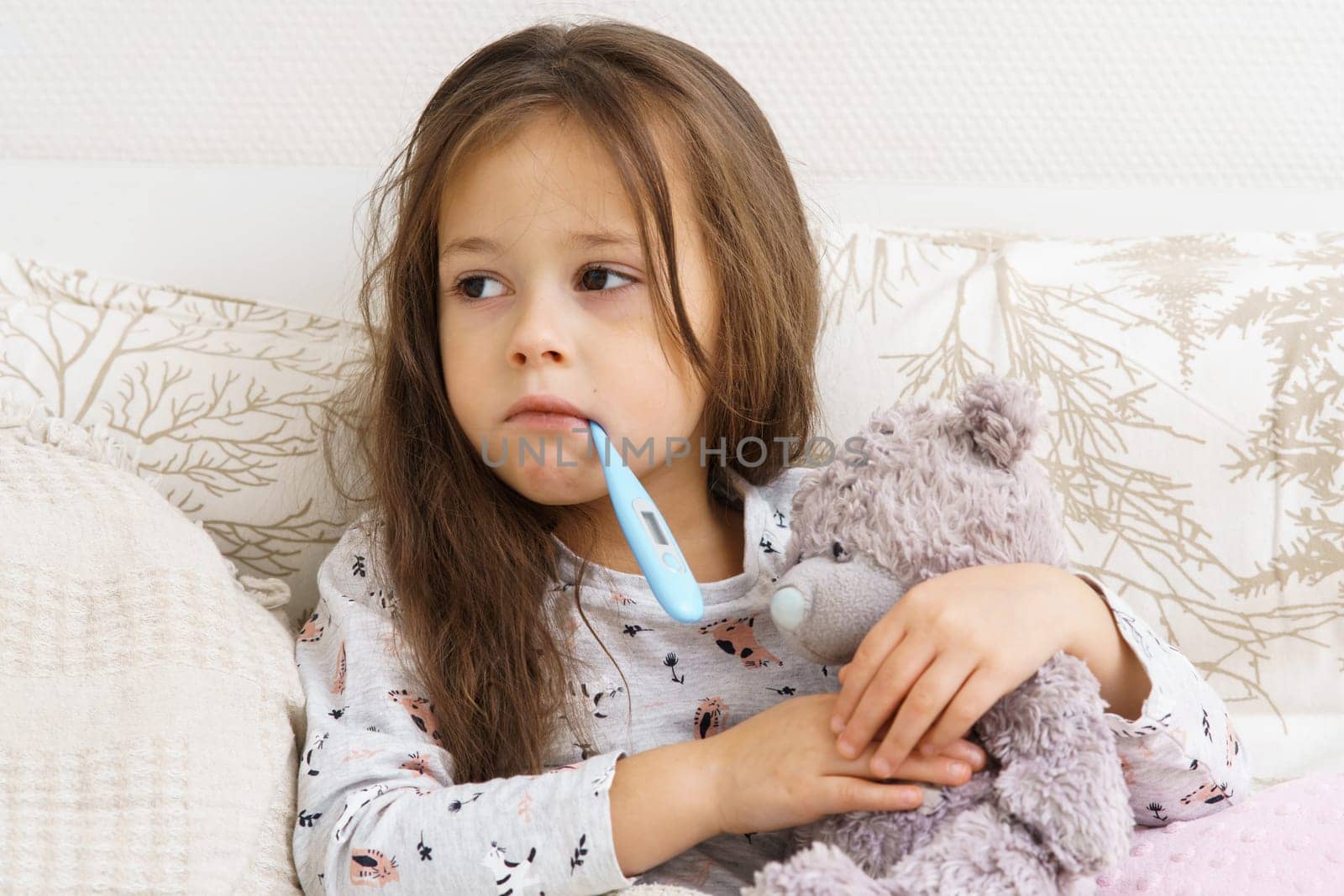 Sick little girl measure temperature with electronic thermometer in mouth and hug teddy bear. Portrait of unhealthy child. Unhappy kid in sleepwear have bed rest. Headache, fever heat, home treatment.