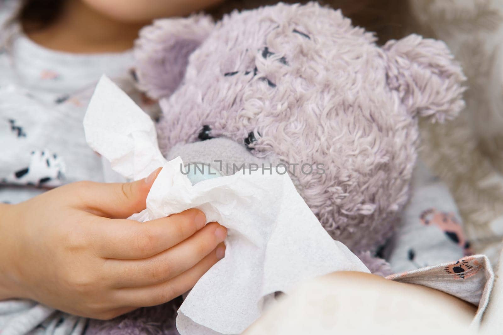 Hand of little girl in sleepwear blow nose of teddy bear with white handkerchief. Portrait closeup. Kid and toy have bed rest. Home patient treatment. Runny nose, cold symptoms, cure.