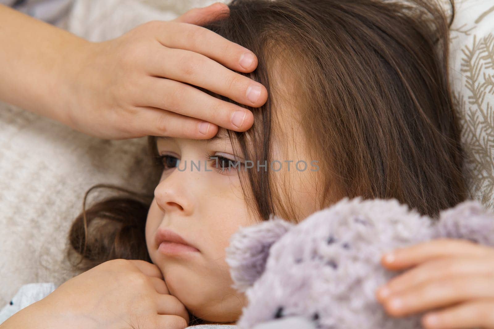 Hand of child touch hot forehead of sick little girl. Portrait closeup of unhealthy child. Kid with teddy bear lie with fever heat. Headache, high temperature. Treatment and bed rest.