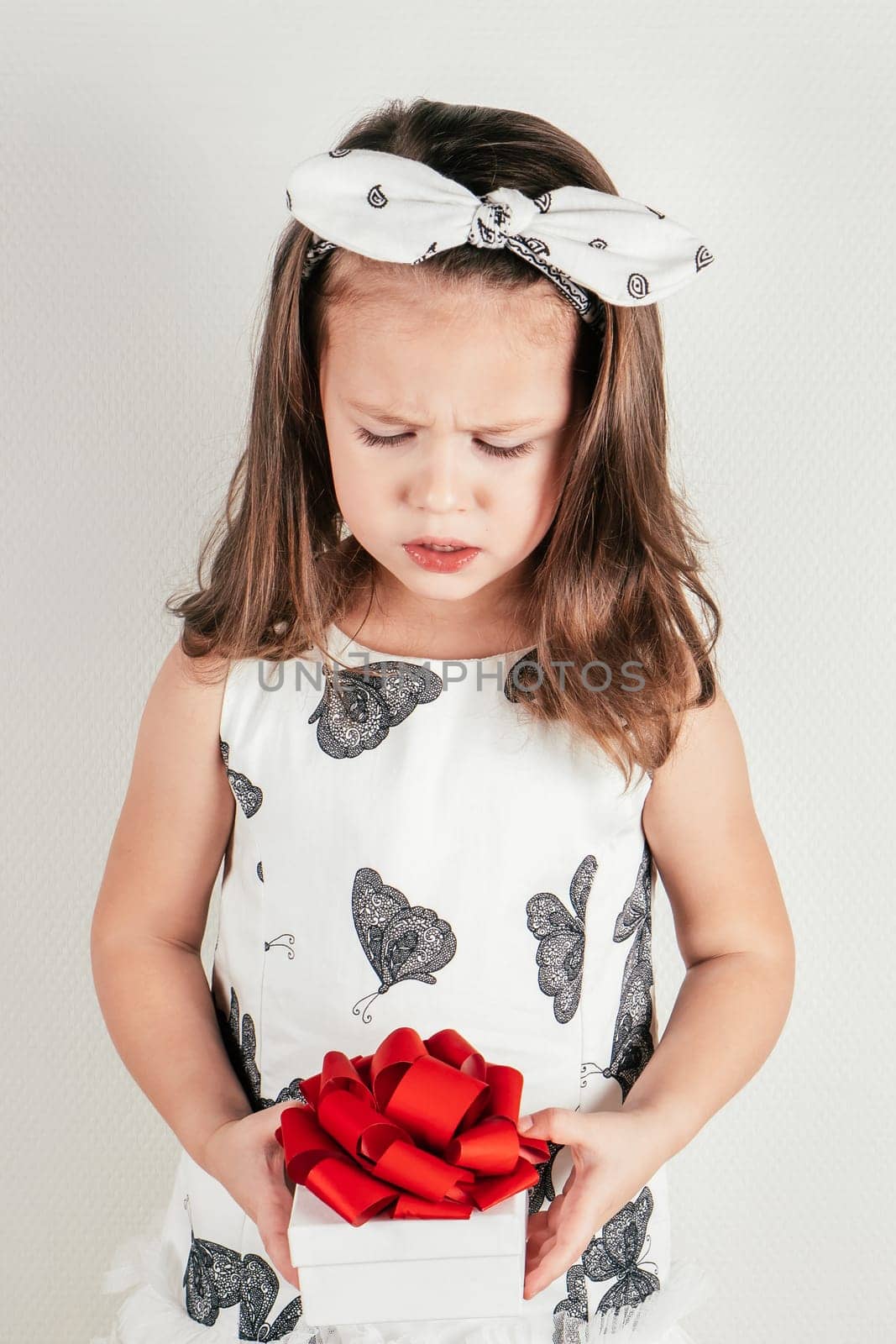 Upset little girl hold beautiful present box with bow looking down. Portrait of unhappy child on white. Pretty kid in festive dress and headband. Saint Valentine's Day concept, holiday of all lovers.