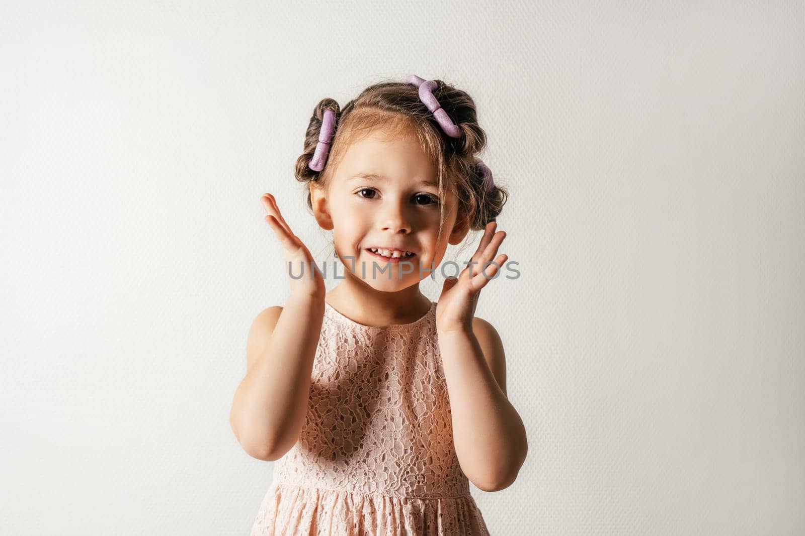 Joyful little girl with hair curlers. Portrait of smiling child on white. Kid in beautiful festive dress prepare hairstyle. Saint Valentine's Day concept, holiday outfit, happy emotions.