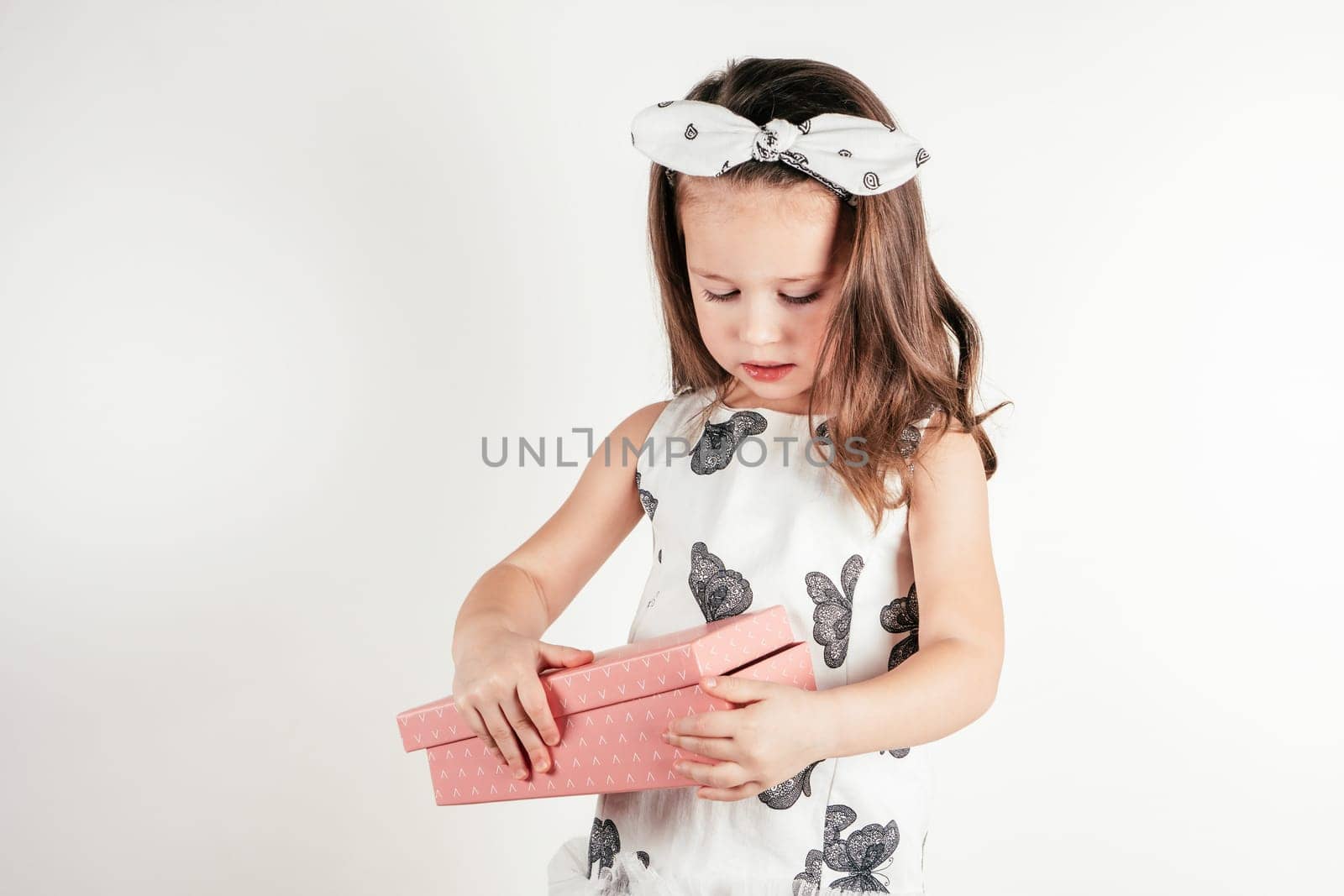 Adorable little girl open pink present box. Portrait of child on white. Pretty kid in festive dress and headband. Gift for holiday. Saint Valentine's Day concept, holiday of all lovers, celebration.