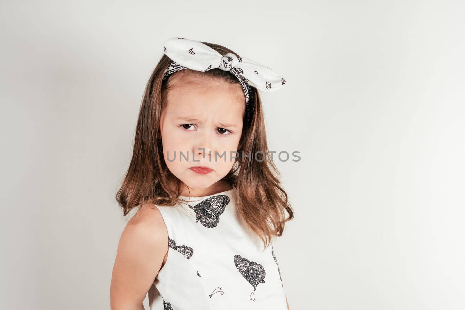 Unhappy little girl with offended facial expression. Portrait of discontent child on white. Kid in beautiful festive dress and headband. Saint Valentine's Day concept, holiday of all lovers.