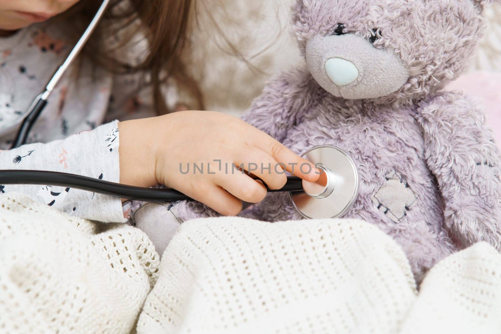 Hand of little girl listening to lung of teddy bear with stethoscope. Portrait of child closeup. Kid and toy sit under blanket and have bed rest. Home patient treatment. Caring doctor.