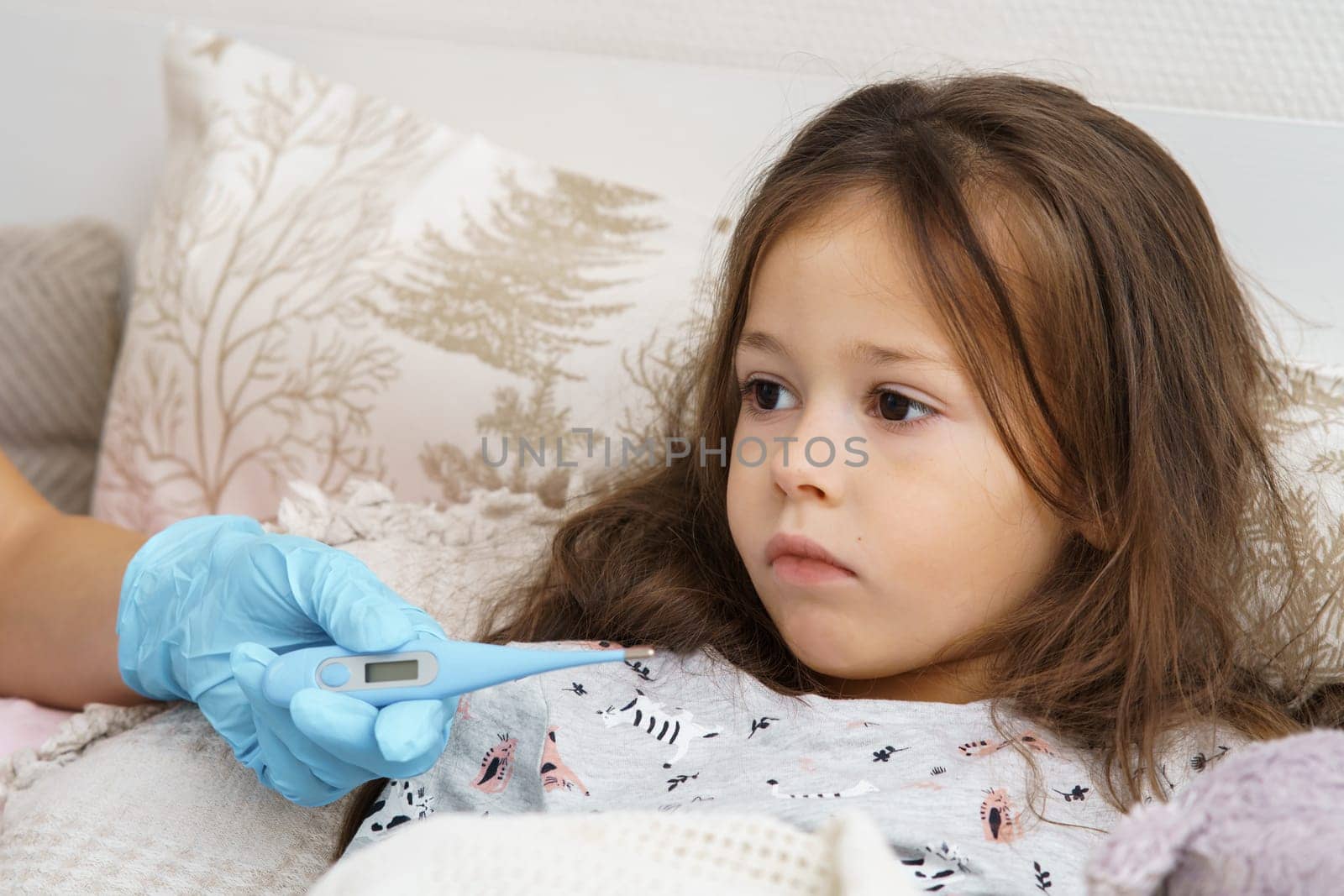 Doctor hand in medical glove measure temperature of sick little girl with electronic thermometer. Portrait of unhealthy child. Kid in sleepwear lie under blanket with fever heat and have bed rest.