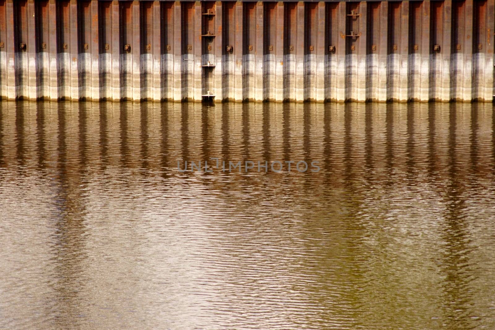 A rusted quay wall and steel river boundary is reflected in the water.