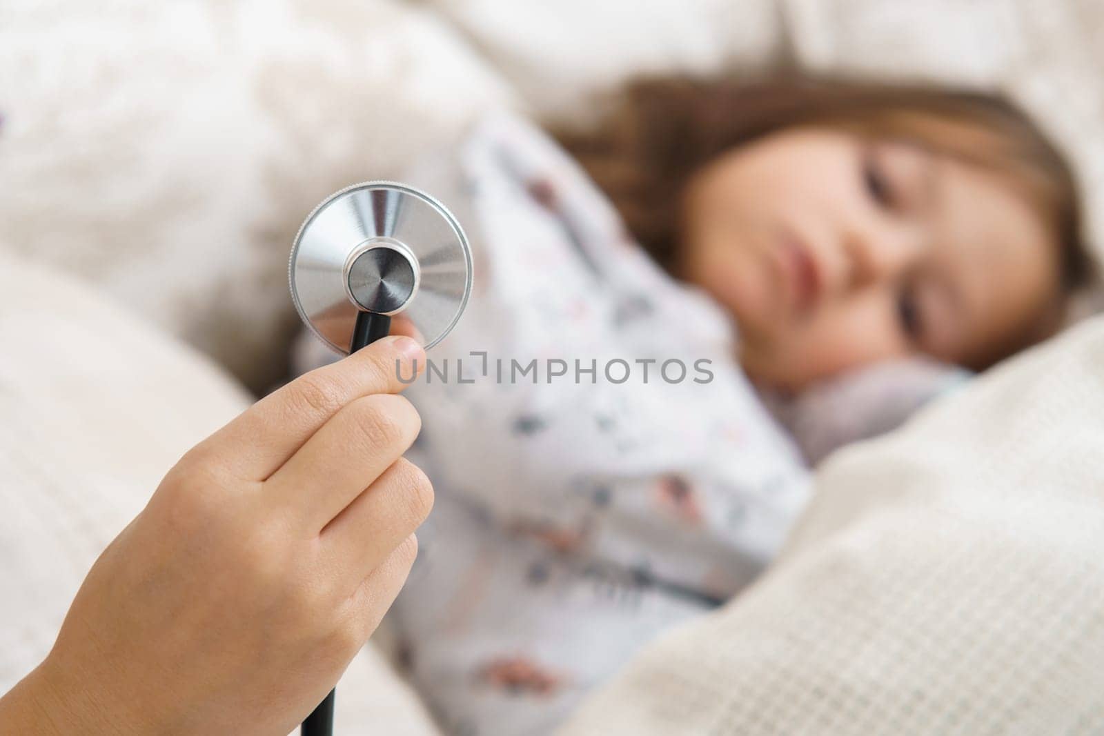 Human hand hold stethoscope for lung listening of little sick girl. Selective focus. Kid in sleepwear lie under blanket and have bed rest. Home patient treatment. Doctor, pediatrician.