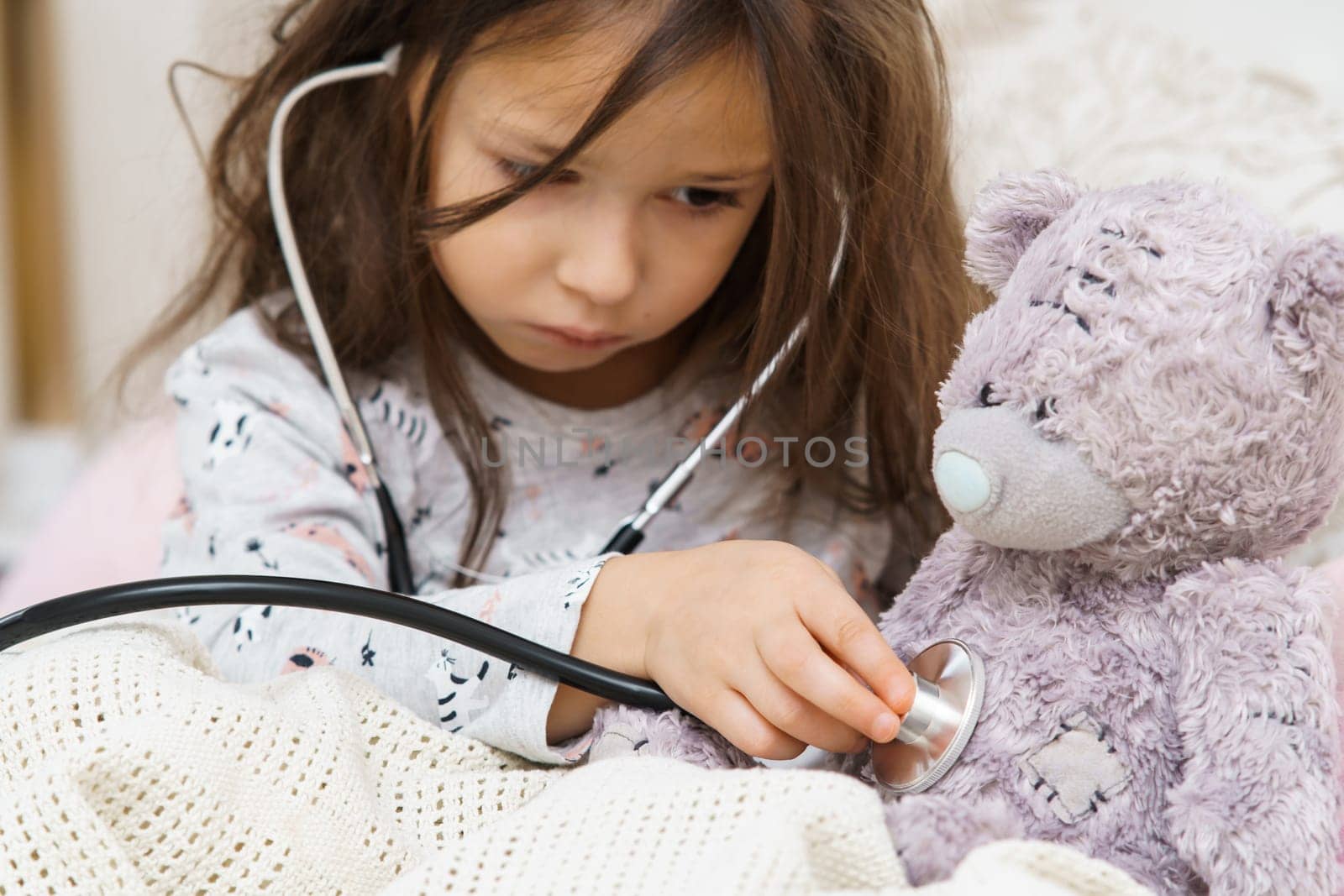 Serious little girl in sleepwear sitting under blanket and listening to lung of teddy bear with stethoscope. Portrait of child closeup. Kid and toy have bed rest. Home patient treatment.