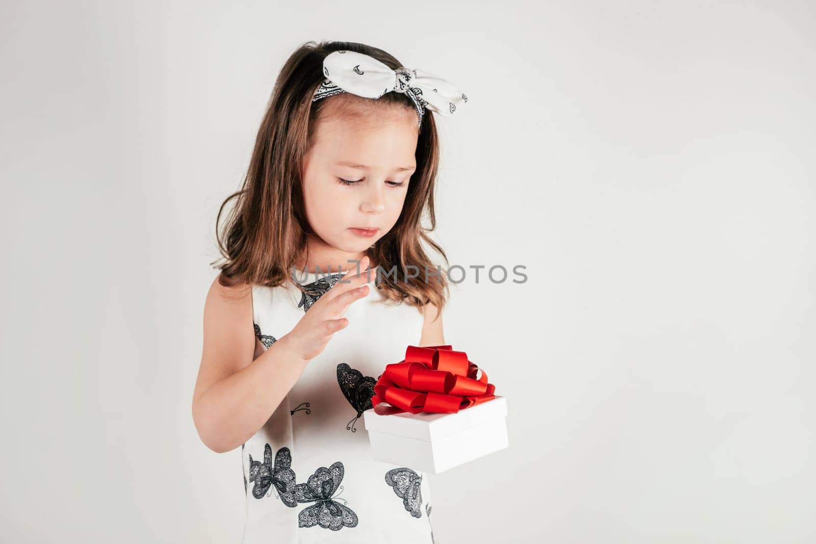 Nice little girl hold beautiful present box with bow looking down. Portrait of child on white. Pretty kid in festive dress and headband. Saint Valentine's Day concept, holiday of all lovers.