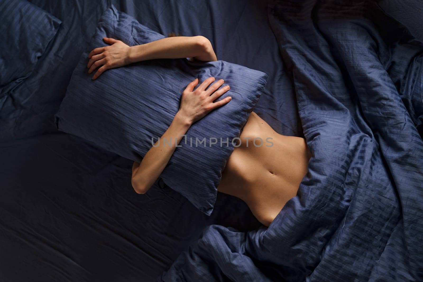 Concept of comfortable rest in bed, lazy weekend and introversion. Woman covers her face with pillow while lying in bed.