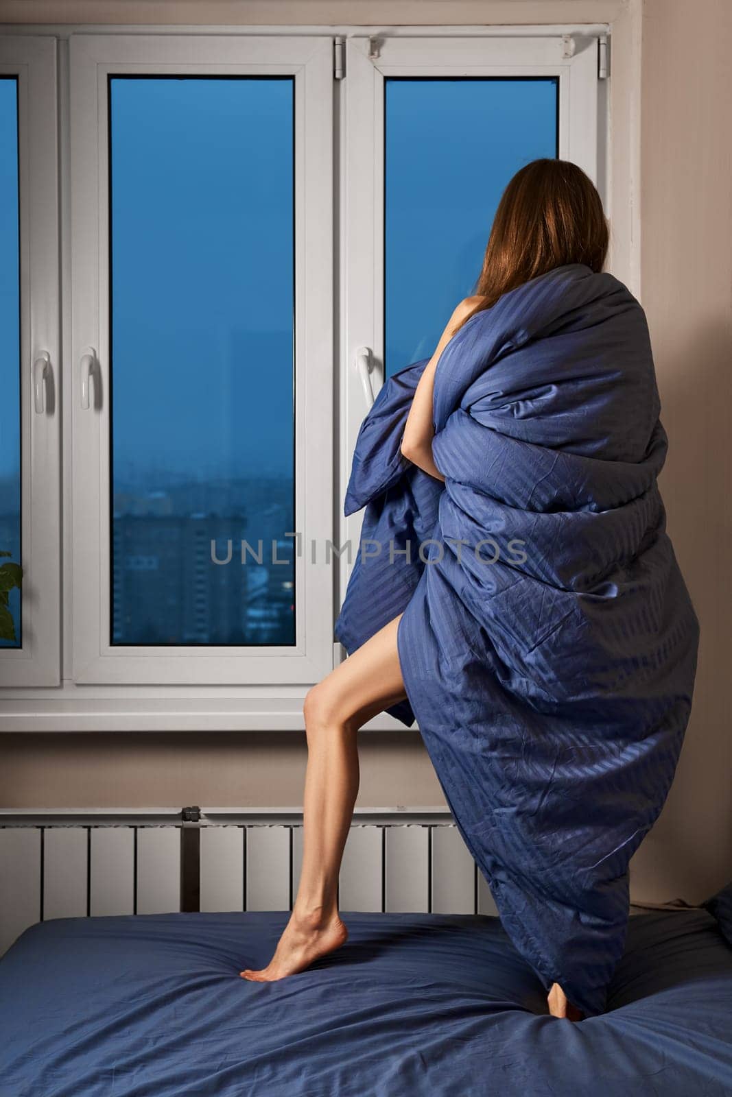Back view of a woman wrapped in soft blanket, standing on the bed and looking out the window