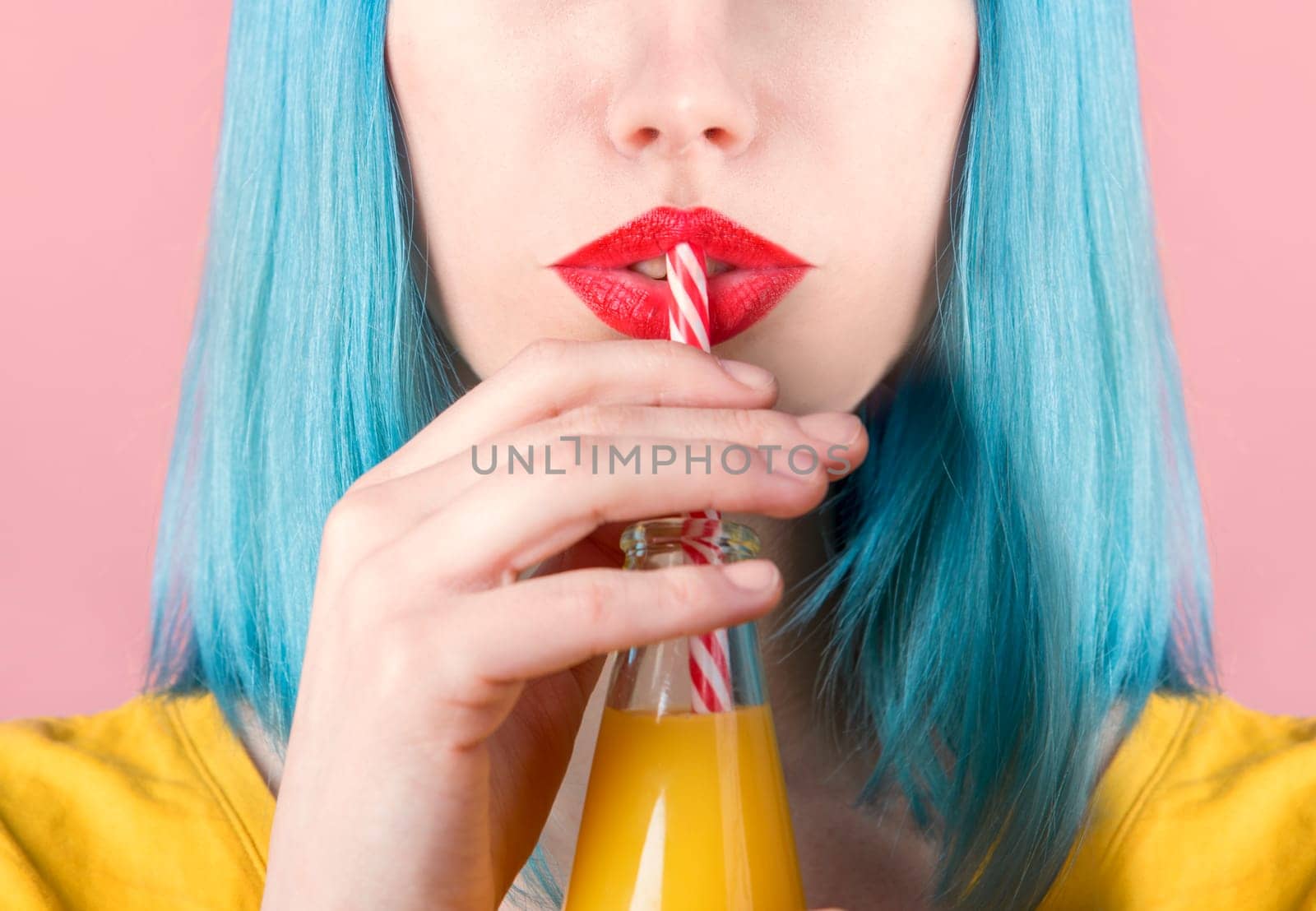 Closeup studio portrait of hipster young woman with red lipstick and blue dyed hair drinking orange juice from retro bottle with straw. Closeup of female mouth drinking summer cocktail. Trendy pastel color background.