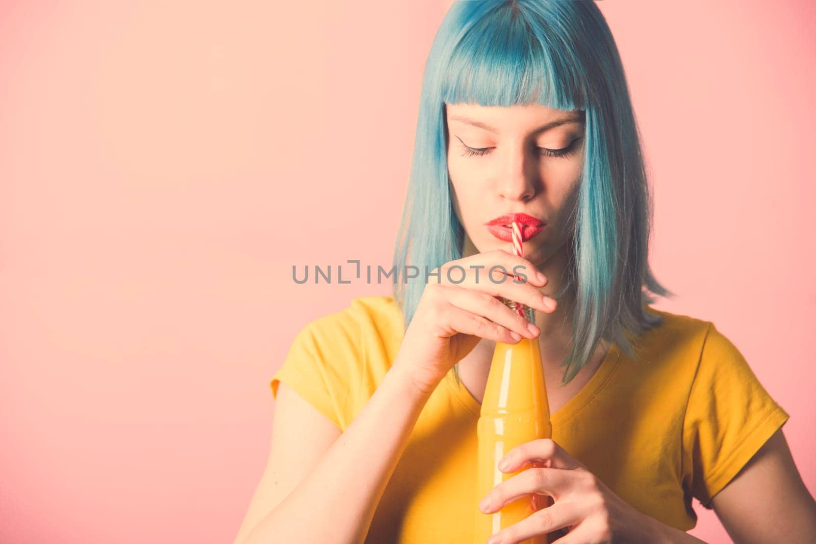Closeup studio portrait of hipster young woman with red lipstick and blue dyed hair drinking orange juice from retro bottle with straw. Closeup of female model holding summer cocktail. Trendy pastel color background.