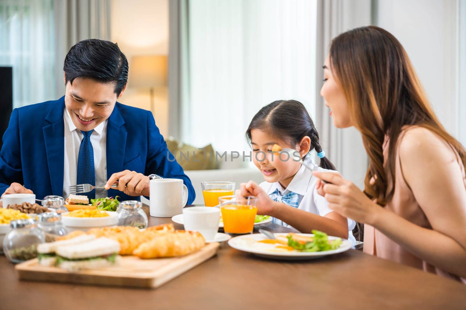 Asian family father, mother with children daughter eating breakfast food on dining table kitchen in mornings together at home before father left for work, happy couple family