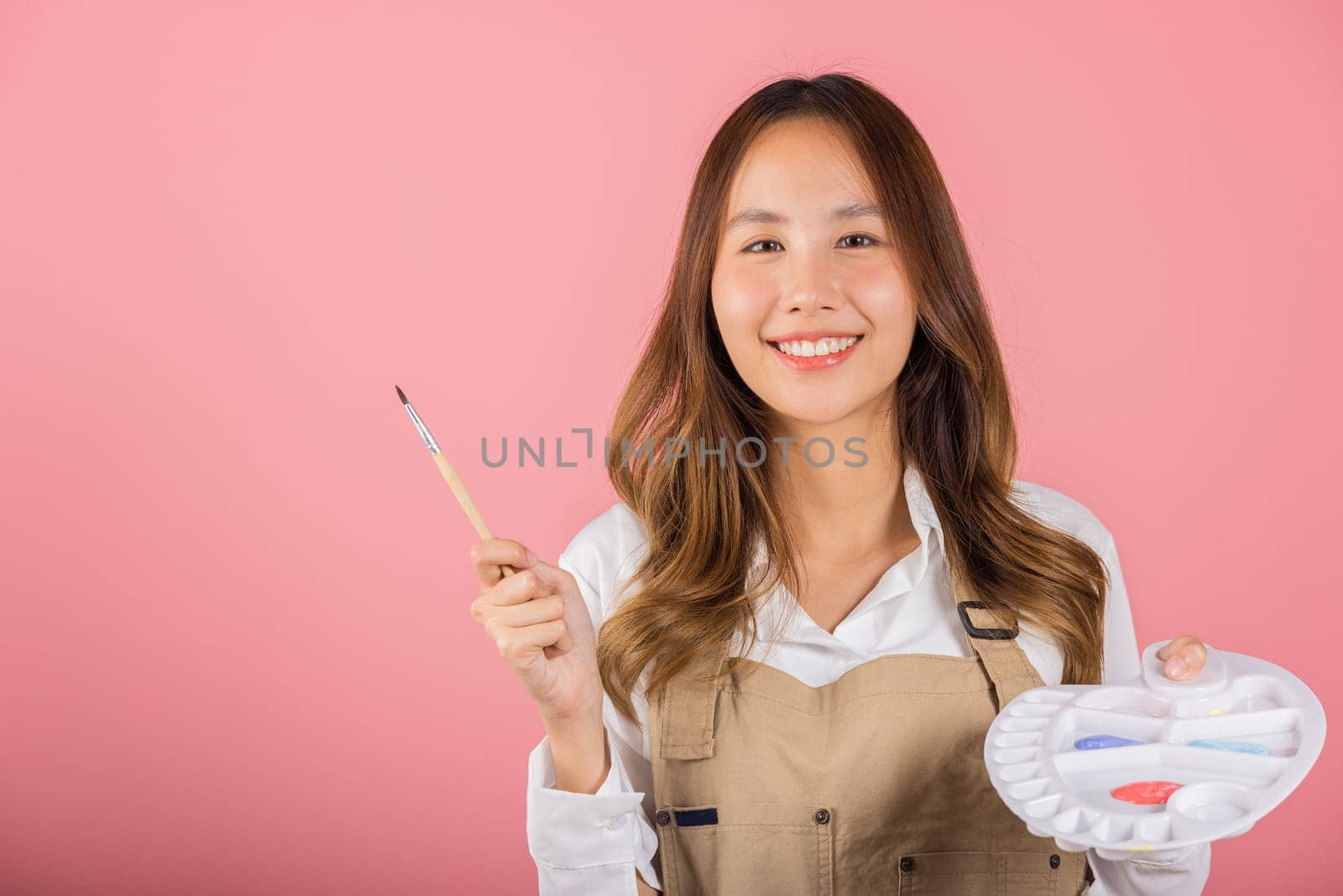 Portrait Asian young woman artist holding brush and paint palette, Happy female painting using paintbrush and palette with colors, studio shot isolated on pink background, Art design workshop