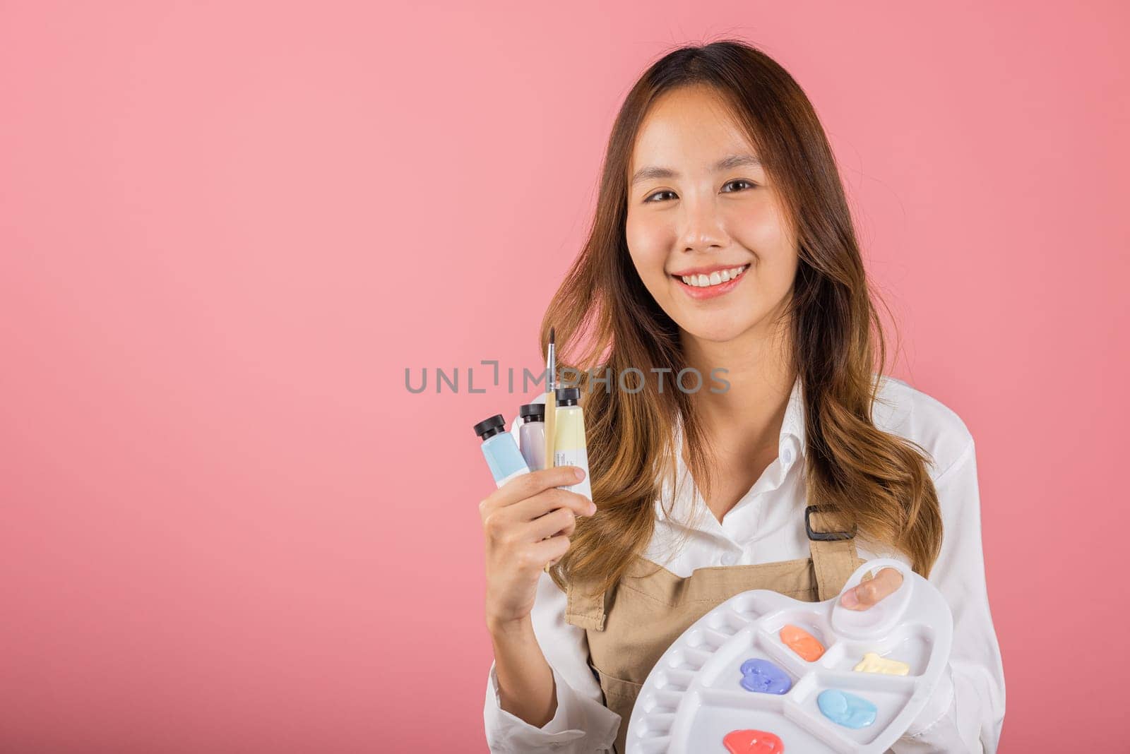 Portrait Asian young woman artist holding brush and paint palette, Happy female painting using paintbrush and palette with colors, studio shot isolated on pink background, Art design workshop