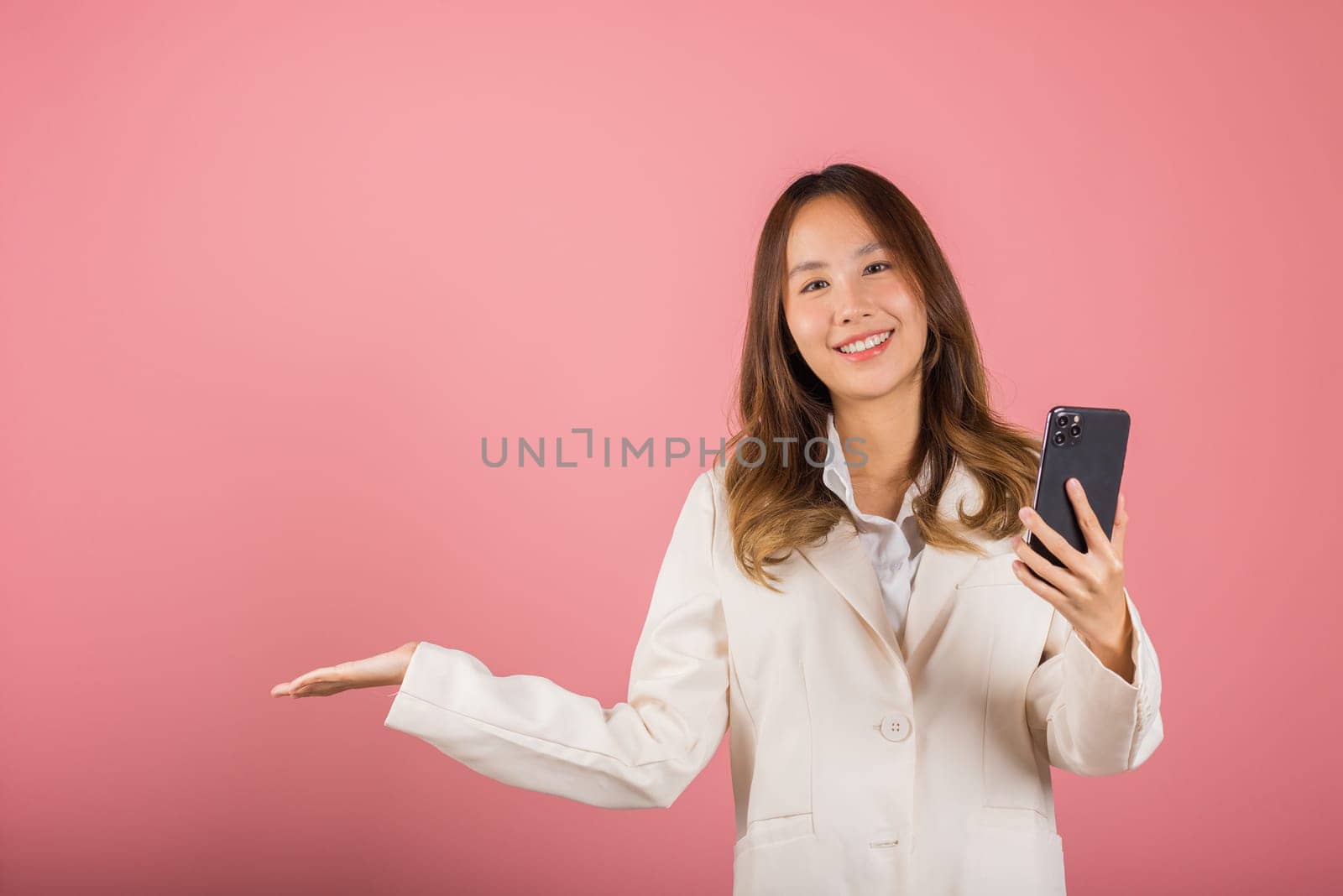 Happy Asian portrait beautiful cute young woman excited holding mobile phone blank screen presenting product with palm of hand at empty, studio shot isolated on pink background, female look camera