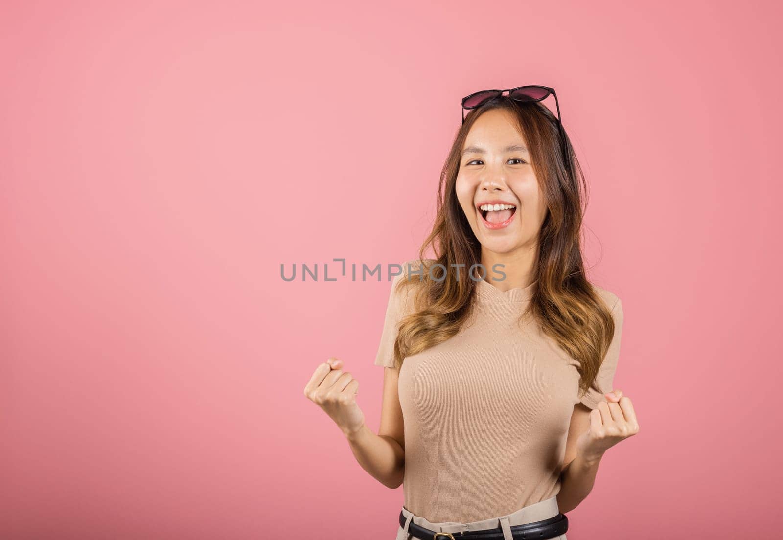 Happy Asian portrait beautiful cute young woman wear glasses makes raised hand up celebrating her winning success gesture, studio shot isolated pink background, Female excited say yes with copy space