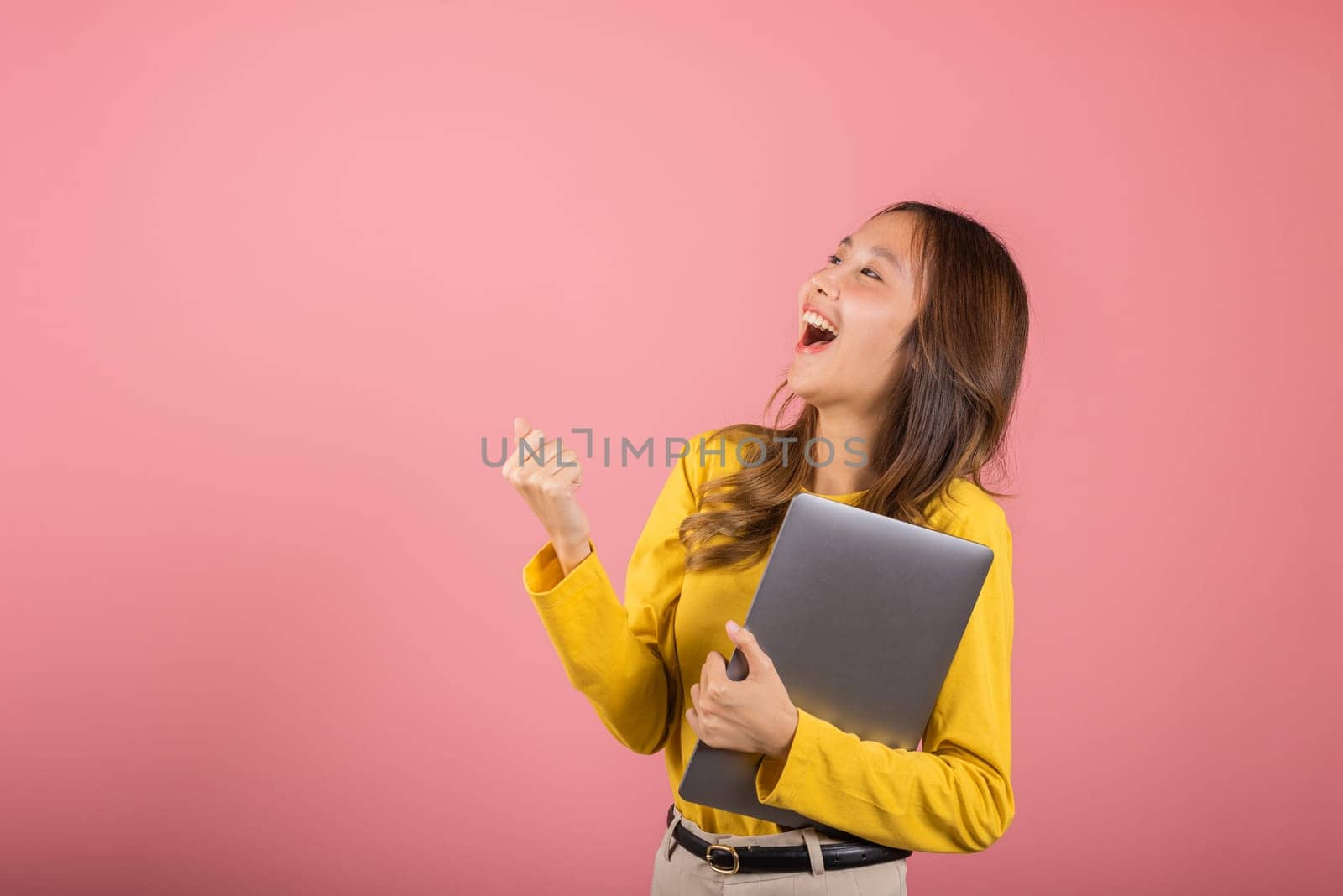 Portrait of happy Asian beautiful young woman confident smiling face holding laptop computer raise hand and say yes excited celebrating success, studio shot isolated on pink background, copy space