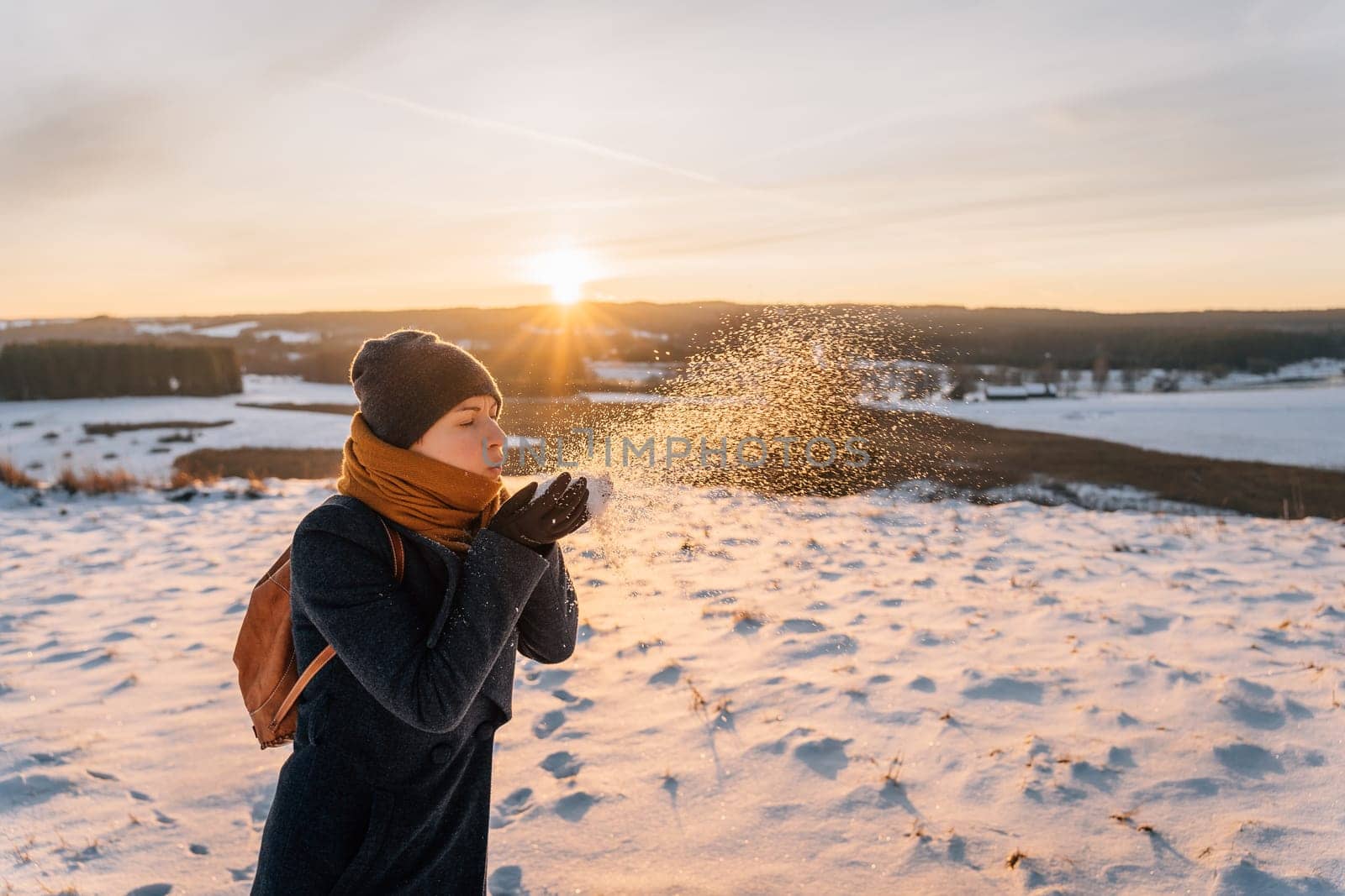 A woman in a coat wrapped in a scarf blows snow off her hands.