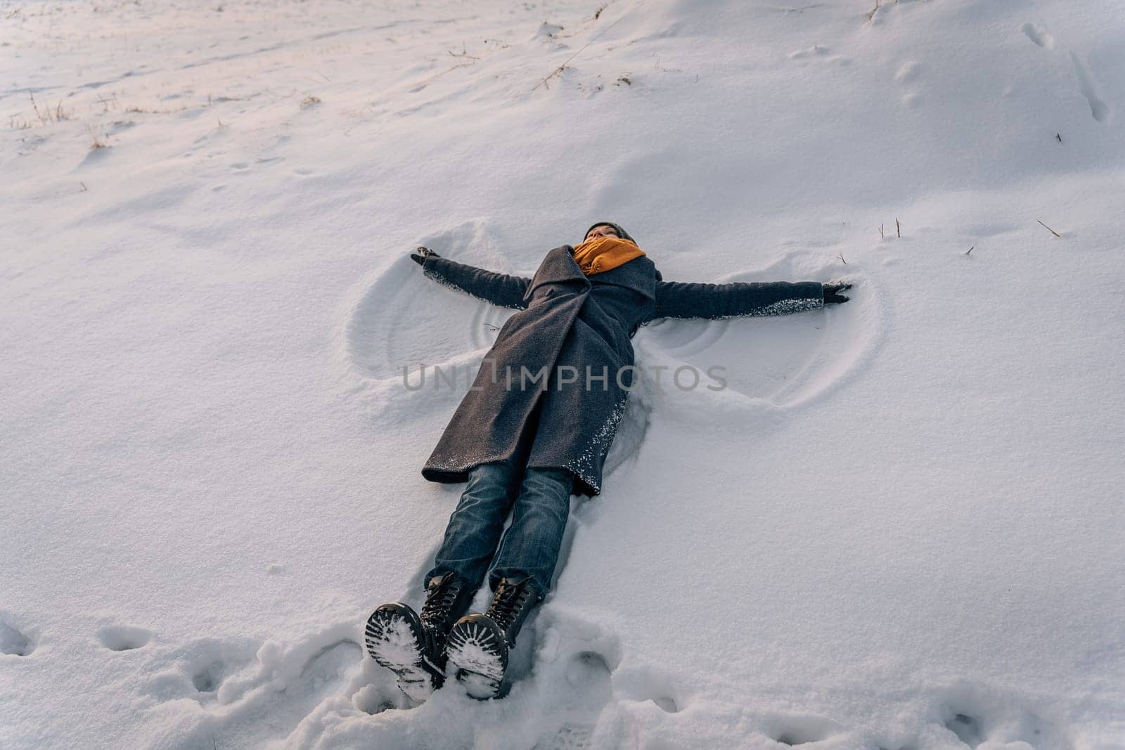 A woman in a coat, scarf and boots lies in a snow-covered field and makes a snow angel.