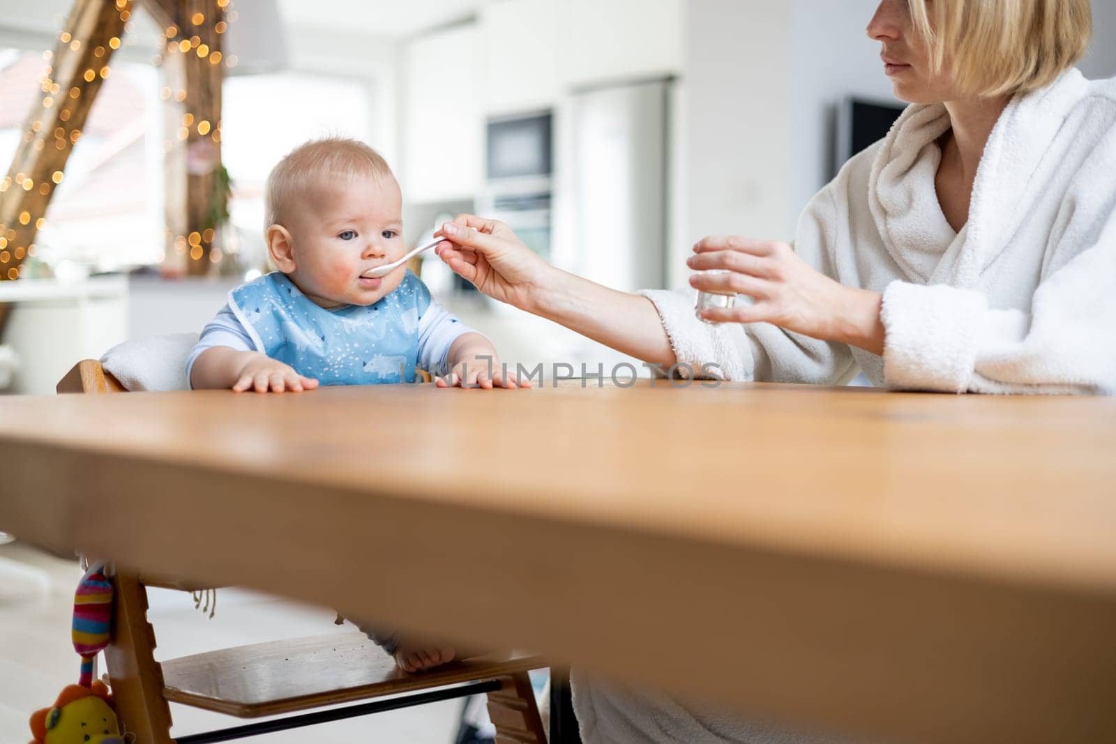 Mother wearing cosy bathrope spoon feeding her baby boy child in baby high chair with fruit puree at dinning table at home. Baby solid food introduction concept.