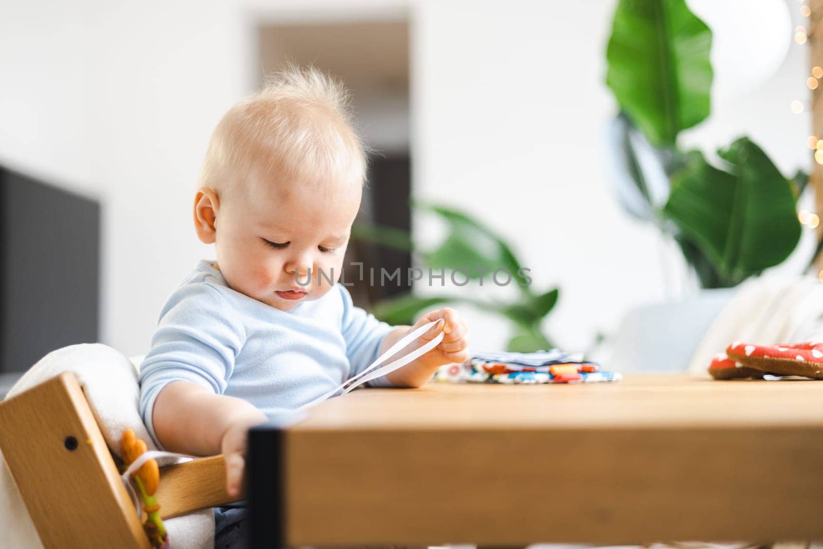 Happy infant sitting at dining table and playing with his toy in traditional scandinavian designer wooden high chair in modern bright atic home. Cute baby playing with toys.