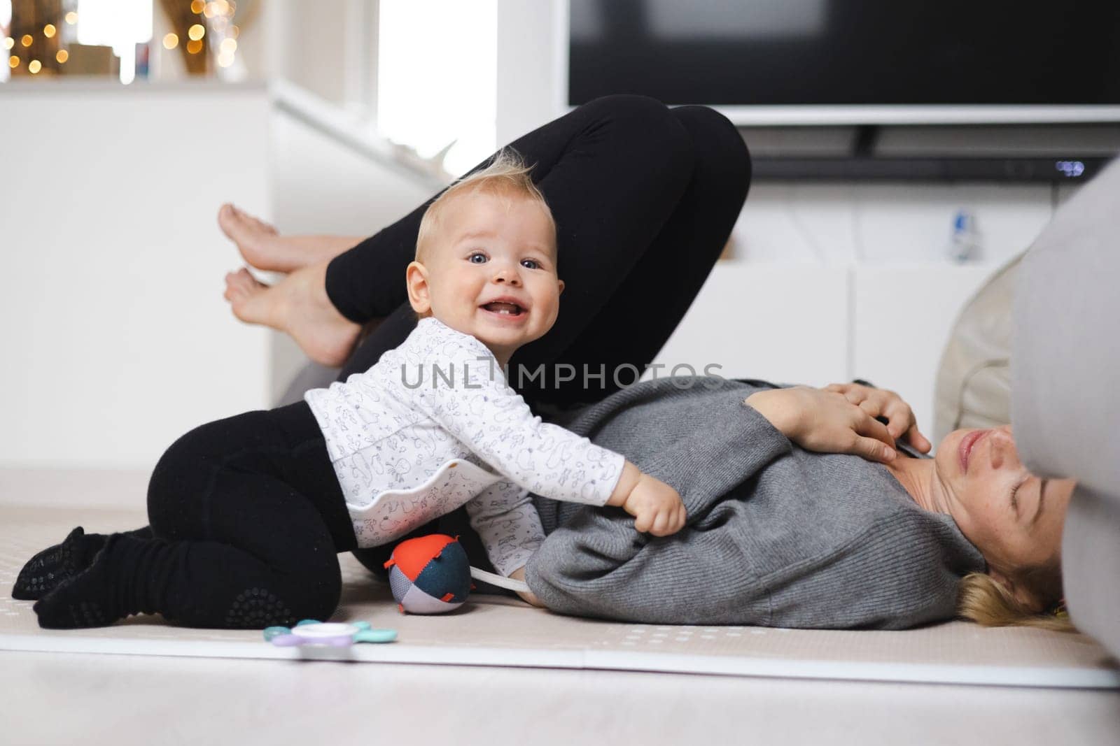 Happy family moments. Mother lying comfortably on children's mat playing with her baby boy watching and suppervising his first steps. Positive human emotions, feelings, joy