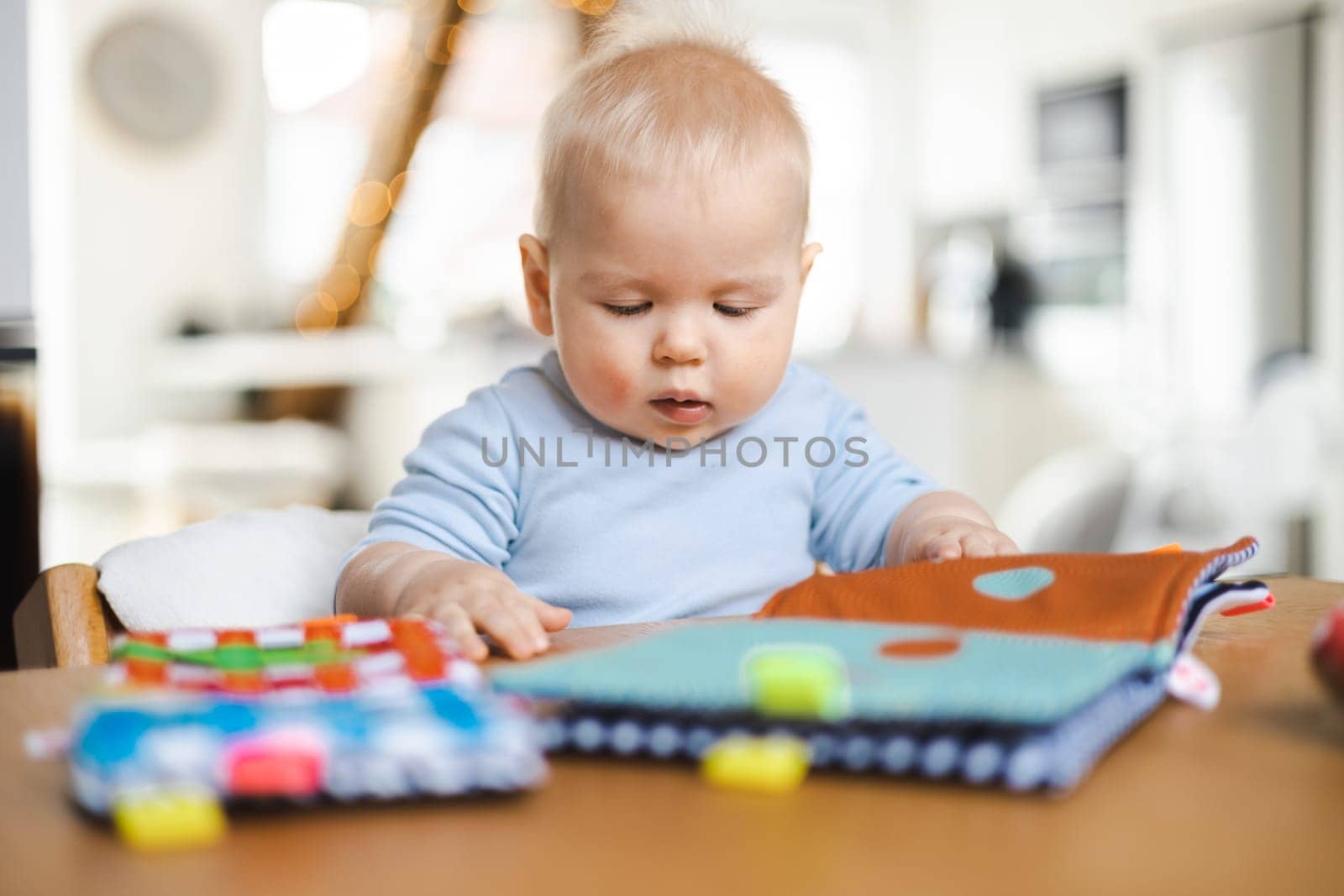 Happy infant sitting at dining table and playing with his toy in traditional scandinavian designer wooden high chair in modern bright atic home. Cute baby playing with toys.