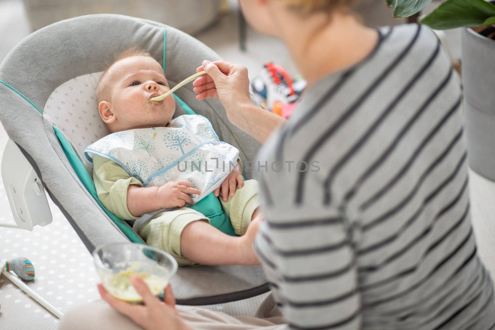 Mother spoon feeding her baby boy child in baby chair with fruit puree. Baby solid food introduction concept