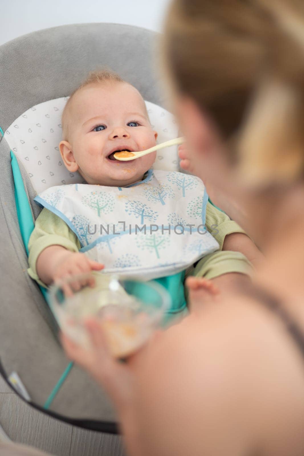 Mother spoon feeding her baby boy child in baby chair with fruit puree. Baby solid food introduction concept