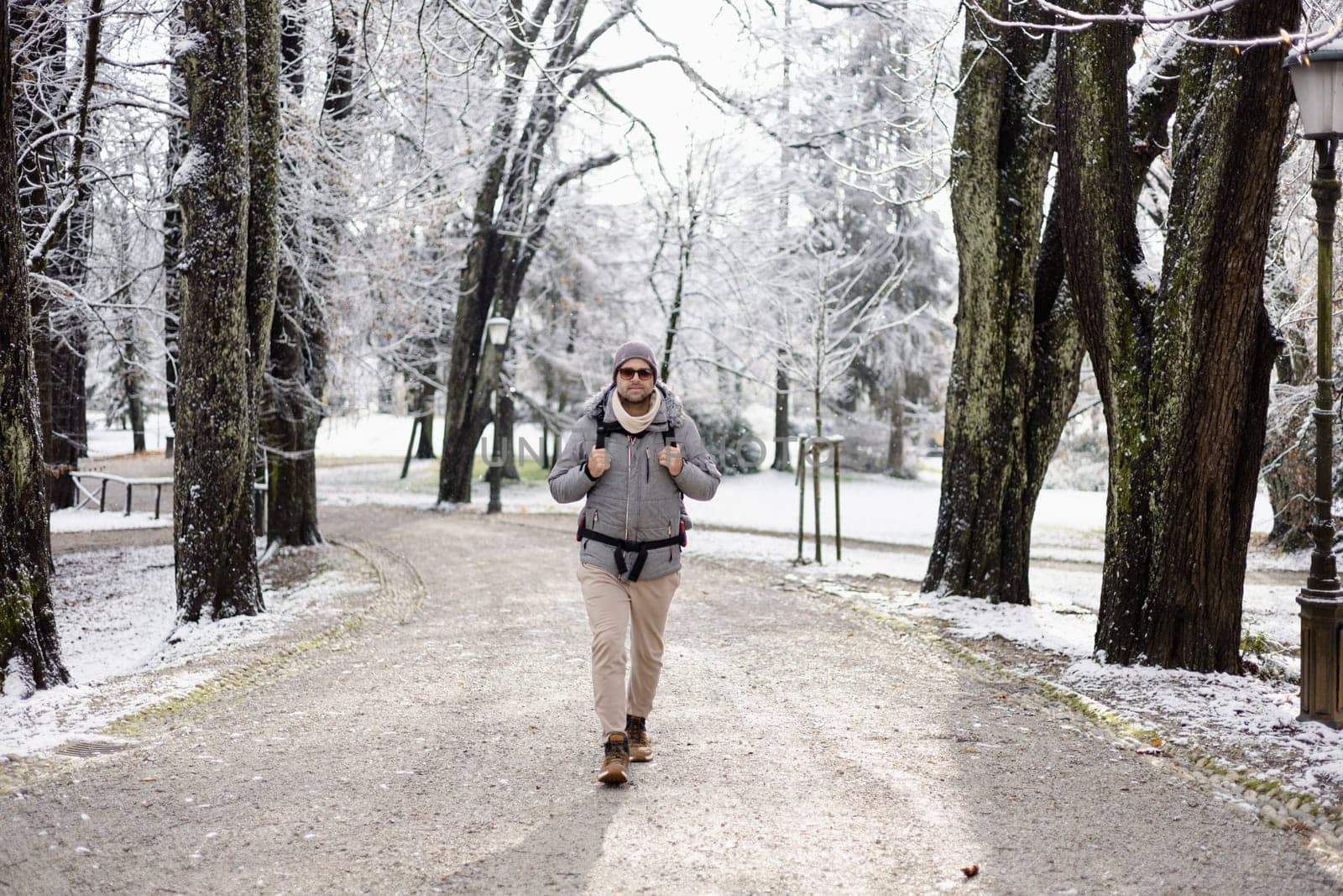 Sporty father carrying his infant son wearing winter jumpsuit and cap in backpack carrier walking in city park in winter.