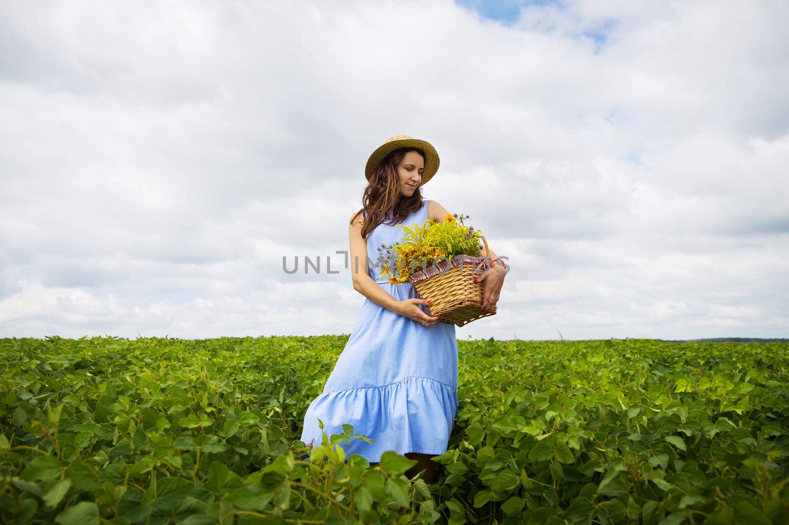 girl in a hat stands on a green field with a basket of flowers.