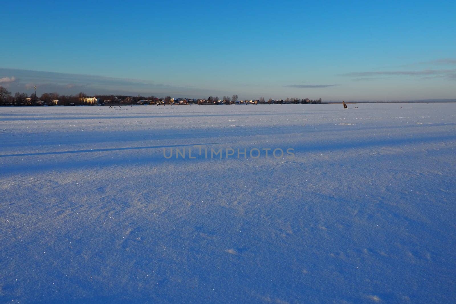Frozen Lake Logmozero, Karelia, Russia. Winter January in nature. Smooth blue ice surface covered with snow. Evening winter sunset in sunny weather. Horizon with coast. Blue sky.