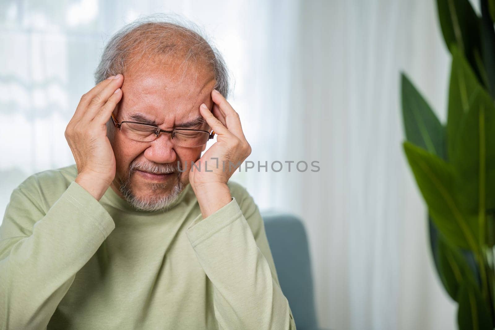 Headache. Sad Asian senior man sitting on sofa feeling hurt and lonely, elderly holds head with hand suffering from migraine headache, Old age health problems, healthcare