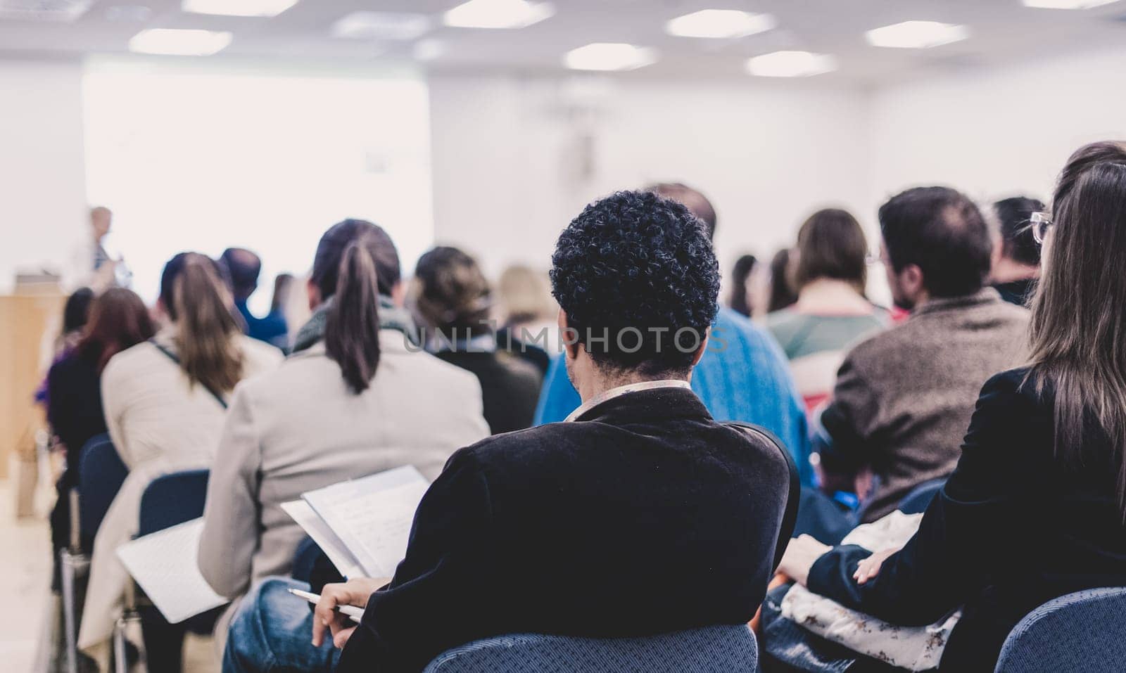 Business and entrepreneurship symposium. Female speaker giving a talk at business meeting. Audience in conference hall. Rear view of unrecognized participants making notes in audience.