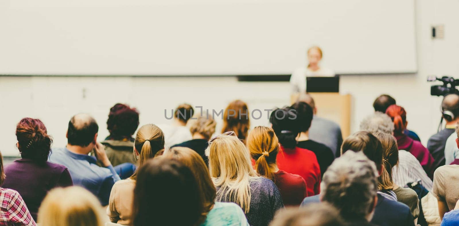 Business and entrepreneurship symposium. Female speaker giving a talk at business meeting. Audience in conference hall. Rear view of unrecognized participant in audience. Copy space on whitescreen.